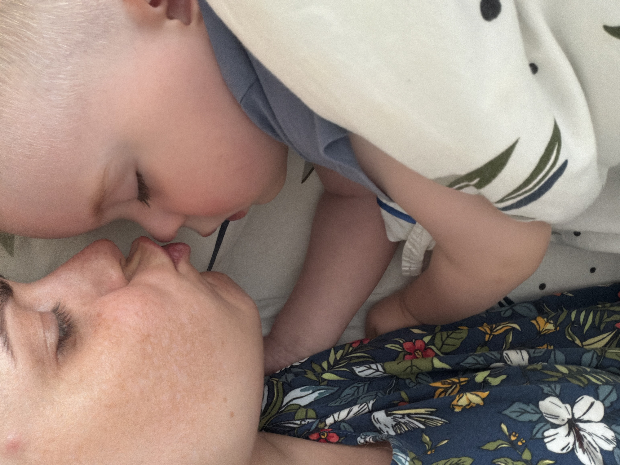 A close-up, slightly angled shot depicts a fair-skinned adult with freckles, lying with eyes closed, and a fair-skinned child with blond hair, also with eyes closed, their faces close together as if about to kiss or touching noses. The adult wears a blue floral patterned garment, while the child is in a light blue shirt. A white blanket with a floral and dot pattern is draped over the child. The background appears to be a bed with a light-colored sheet. The location is not discernible from the image.