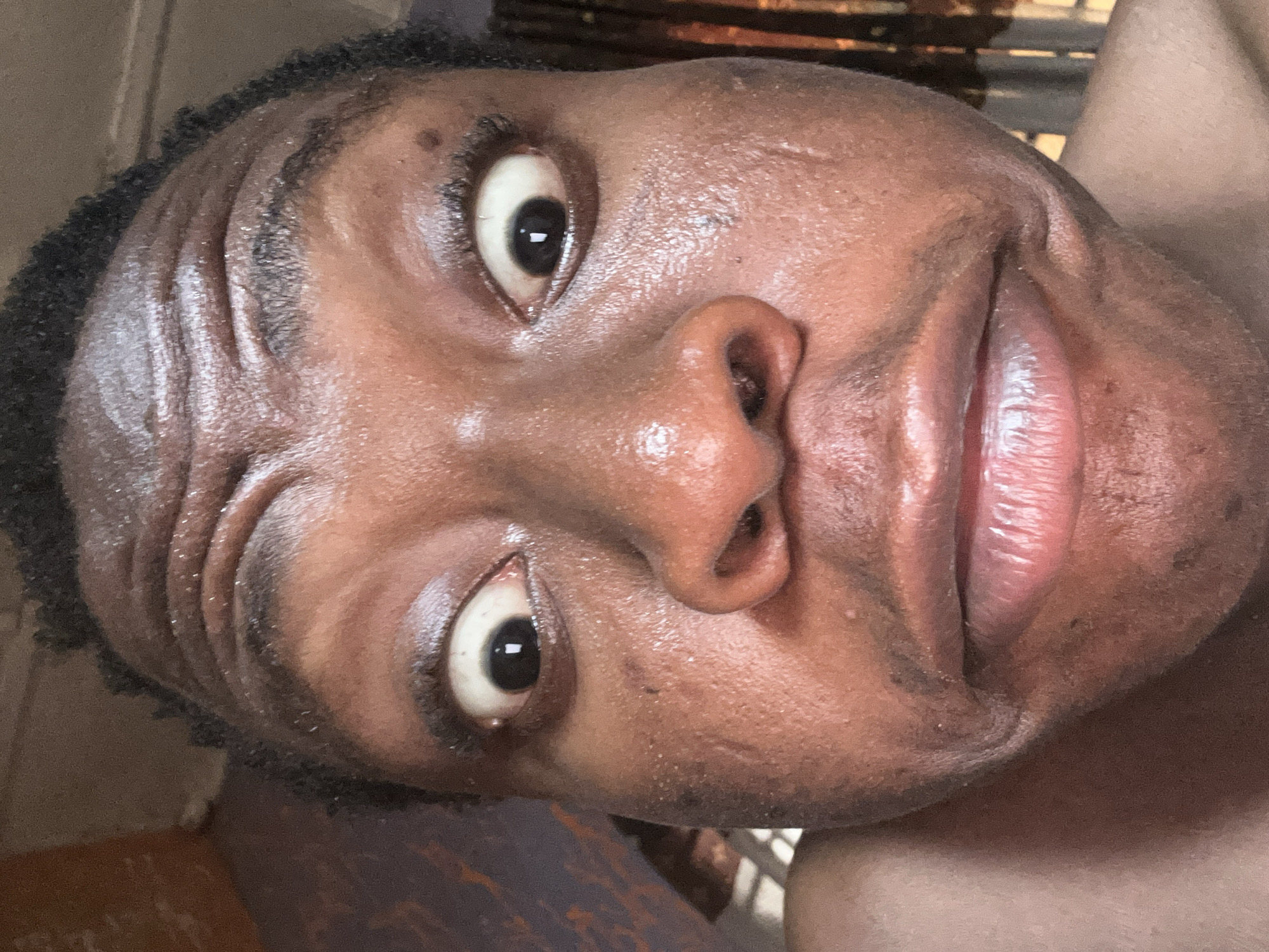 A close-up, slightly angled portrait shows a Black male with dark hair, eyes, and skin. His face appears shiny, and his lips are slightly parted. The background is blurred and indistinct.
