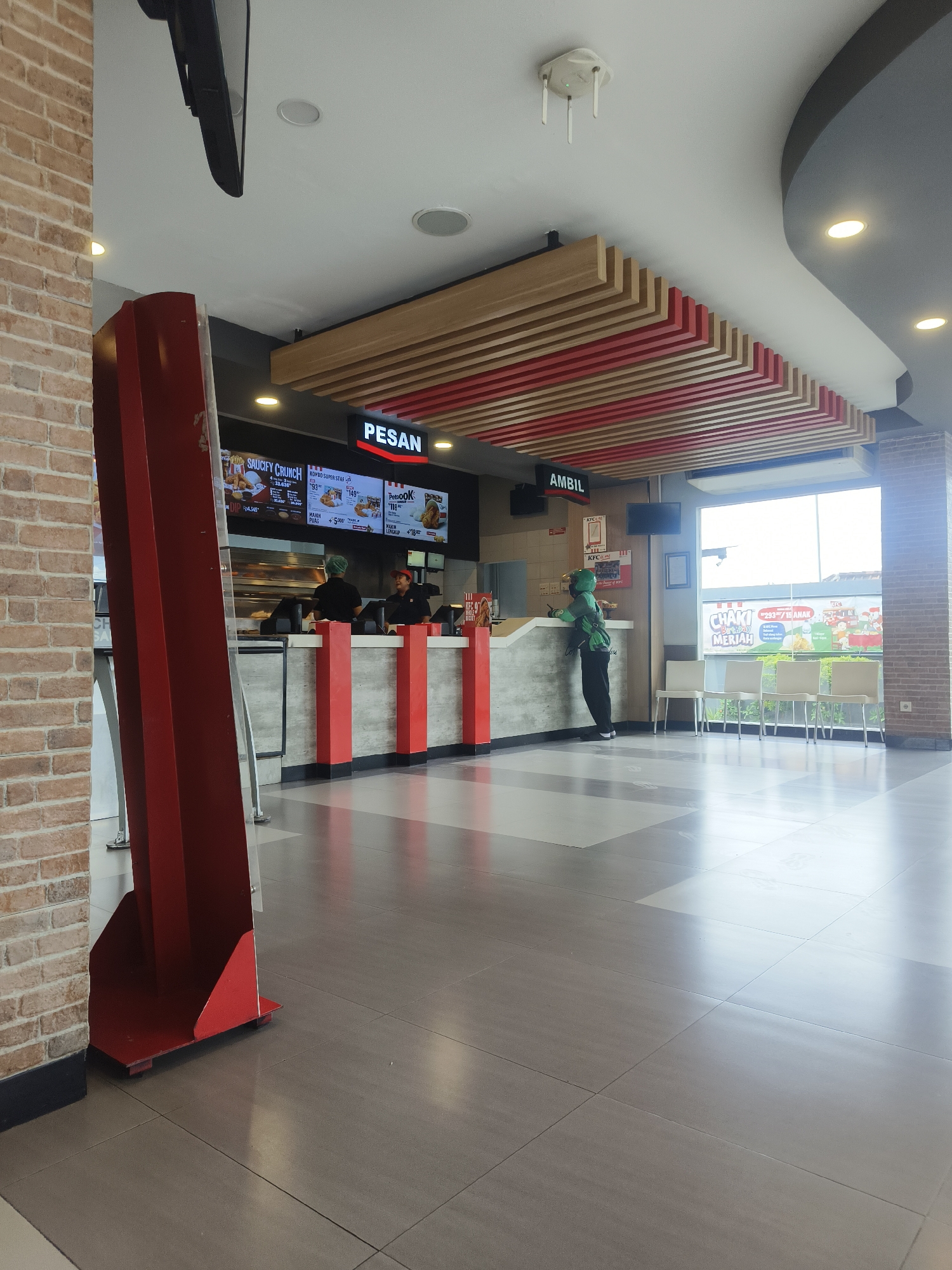 The image shows the interior of a KFC restaurant in Denpasar, Indonesia. A counter with two employees in black uniforms is visible, serving customers. A customer in a green uniform and helmet is at the pick-up counter. The restaurant features a modern design with a suspended wooden ceiling and tiled floors. A large window looks out onto an outdoor area with chairs and tables. The overall atmosphere is clean and well-lit.