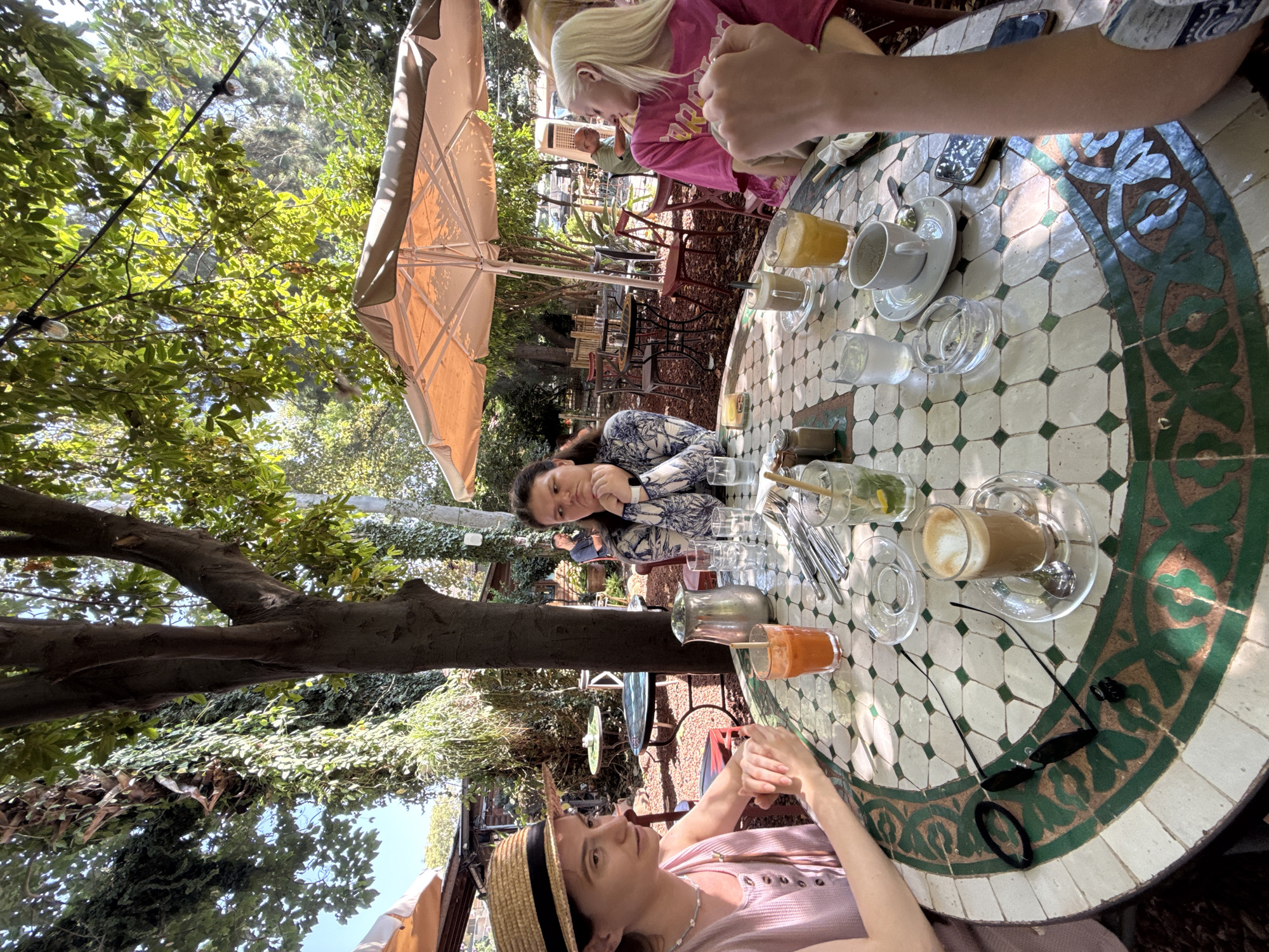 Four individuals are seated around a table in an outdoor cafe in Mevoot HaHermon Regional Council, Israel. The table is circular and decorated with a mosaic pattern of white, green, and brown tiles. On the table are several drinks including a glass of orange juice, a coffee with froth, and a glass with ice and lemon. Cutlery, glasses of water, a sugar dispenser, and an ashtray are also present.
On the left, a woman wearing a straw hat and a pink top is seated facing the camera, looking to the side with her hands clasped on the table. To her right, a person with dark hair, wearing a blue and white patterned shirt, is looking down with their chin resting on their hand. Across from them, a person with blonde hair, wearing a bright pink t-shirt, is partially visible, their arm reaching towards the center of the table. A fourth person's arm is in the foreground on the right.
The cafe setting is lush and green, with trees and foliage surrounding the seating area. Sunlight filters through the leaves, creating dappled light on the table and ground. Umbrellas provide shade over some of the tables. In the background, other tables, chairs, and patrons are visible, as well as some buildings and trees. The overall atmosphere appears relaxed and casual, indicative of a pleasant day at an outdoor eatery.