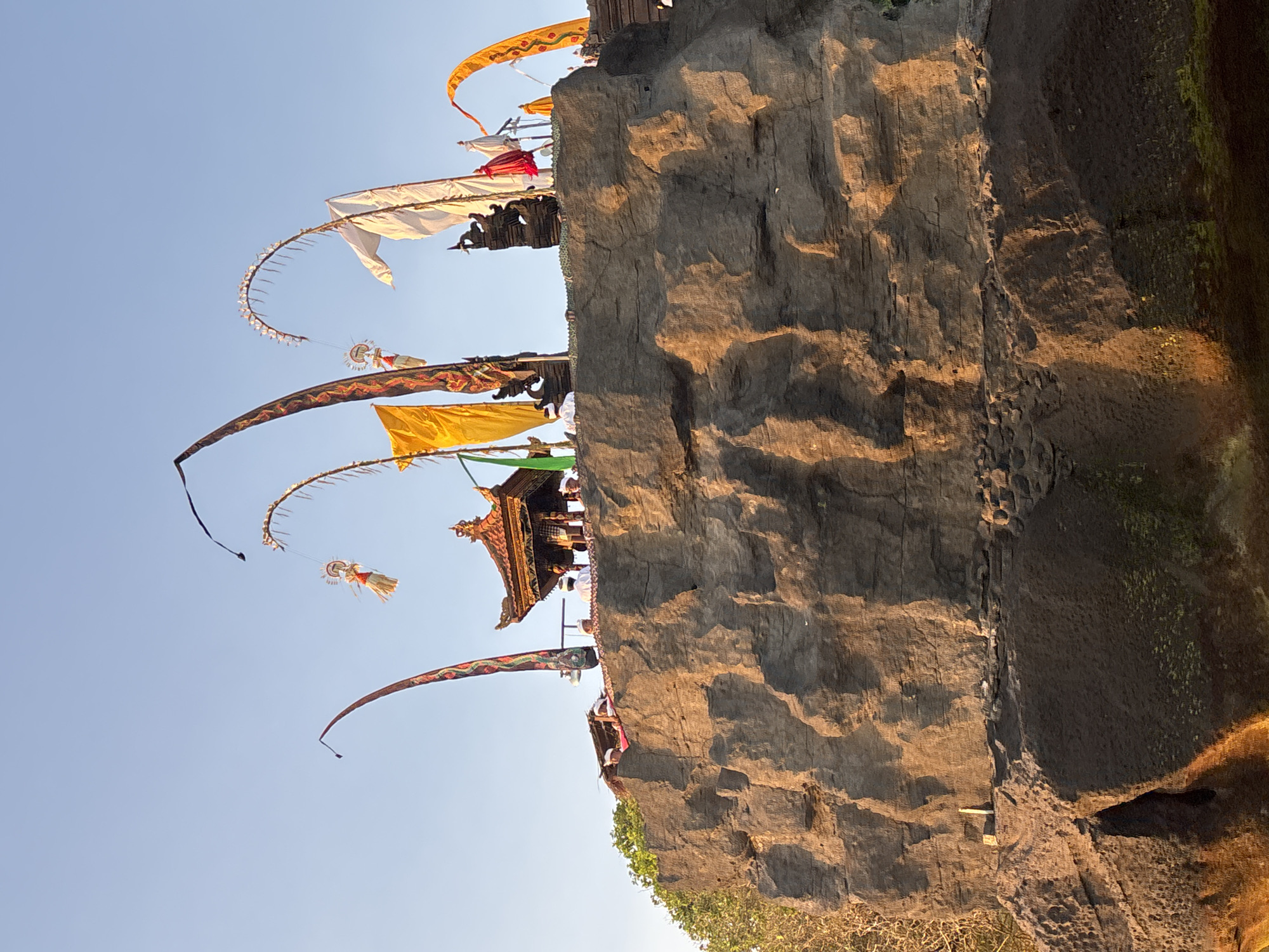 The image depicts a cliffside temple in Kerambitan, Indonesia, set against a clear, blue sky. A large, textured rock face forms a significant portion of the composition, with a temple structure visible on top of the cliff. Colorful Balinese flags and pennants are arrayed along the cliff's edge and extend into the sky, including the distinctive 'penjor' bamboo poles with decorations. People, presumably worshippers or temple attendants, can be seen near the temple structure. The lighting indicates either sunrise or sunset, casting warm shadows on the rock face. At the bottom of the image, the edge of vegetation is visible.