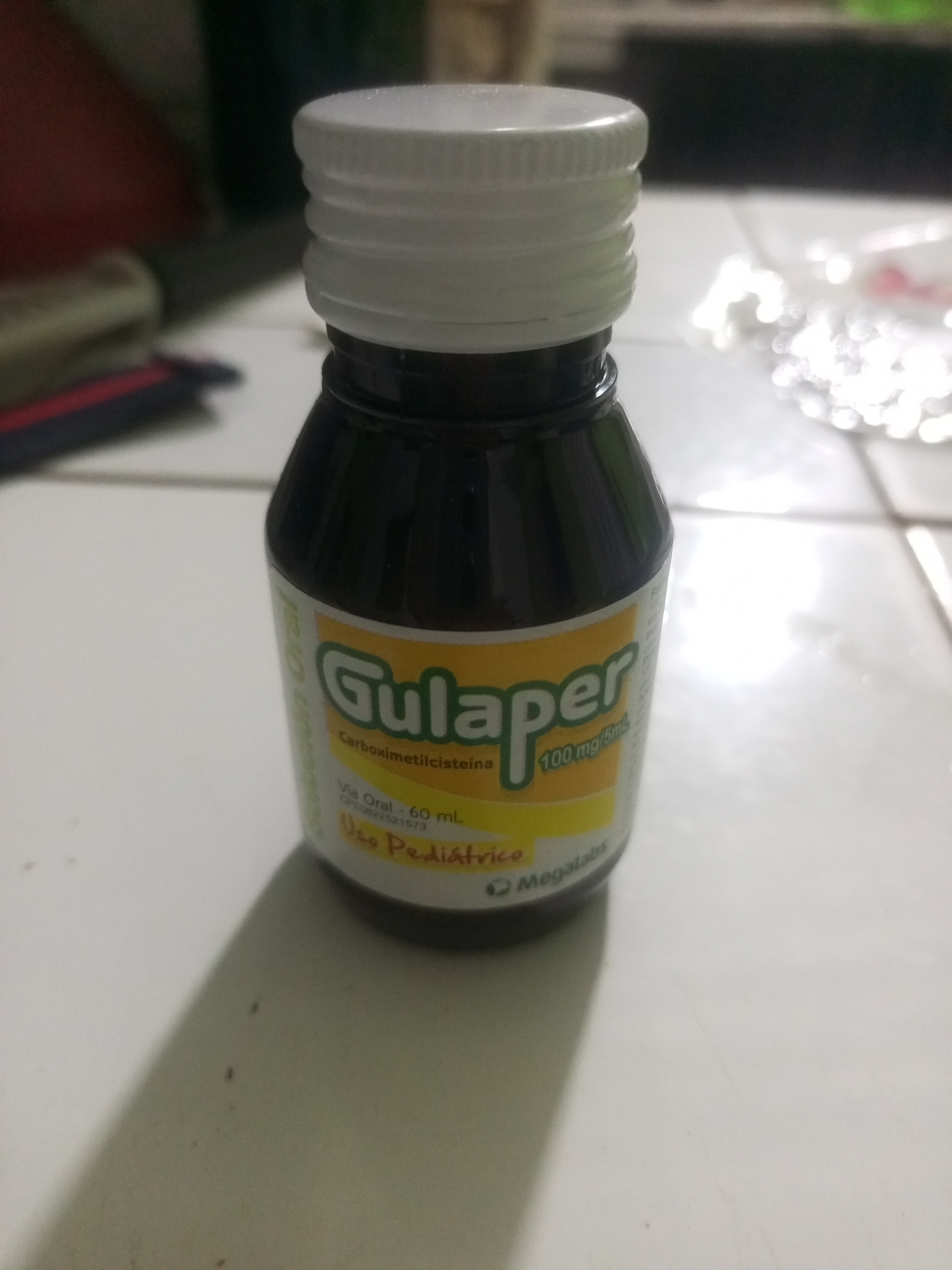 A dark glass bottle of "Gulaper" medication with a white screw-on cap sits on a tiled surface. The bottle has a yellow label with green lettering that reads "Gulaper," and below it, in smaller text, "Carboximetilcisteina 100 mg/5mL." Further down the label, it says "Via Oral - 60 mL" and "Uso Pediátrico," along with a company logo that appears to be "Megalabs." The bottle is casting a shadow on the tiled surface. The background is blurred and out of focus, showing hints of a dark surface and some light-colored, potentially reflective objects. There is no indication of the location being in the city of La Tagua, Venezuela, or any other specific geographical location. The scene appears to be indoors, possibly on a table or countertop.