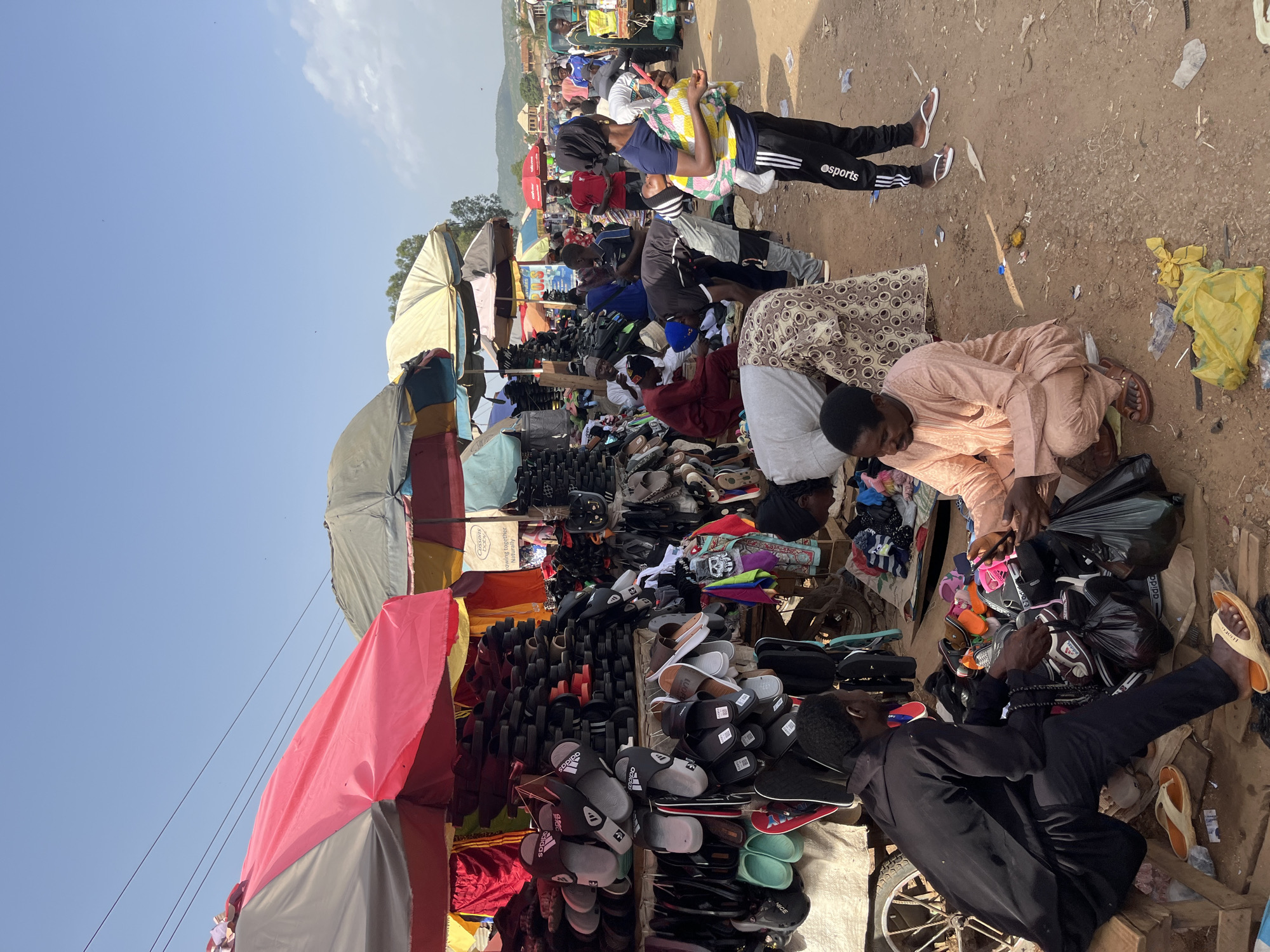 A street market scene in Rafin Par, Nigeria, features numerous stalls adorned with colorful umbrellas, displaying a variety of goods, primarily footwear. Dominating the foreground is a vendor in a black tunic and trousers, seated and inspecting footwear, with an array of sandals and flip-flops displayed on shelves and in piles around him. To his right, another individual, wearing a peach-colored traditional outfit, is also engaged with the merchandise, holding a mobile phone and a plastic bag. The ground is dirt and littered with debris, including plastic bags and wrappers. In the background, other market-goers and vendors are visible amidst the stalls, with lush greenery and residential structures in the distance, suggesting a bustling commercial area within the town. The sky is clear and blue.