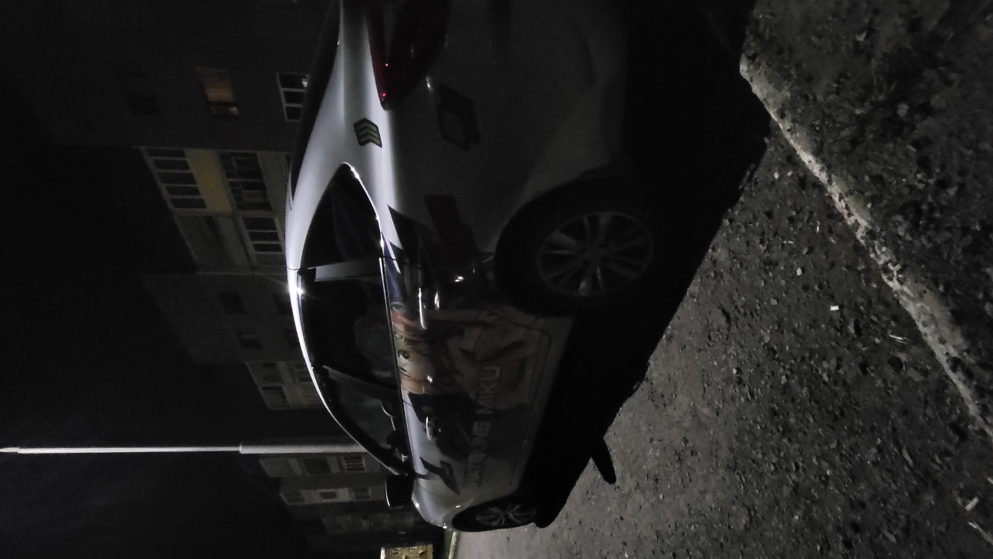A car with a "Hatsune Miku" decal on the side is parked on a street in Марковский, Russia. The background shows buildings and a streetlight. It is dark. The car is parked on a paved road next to a curb.