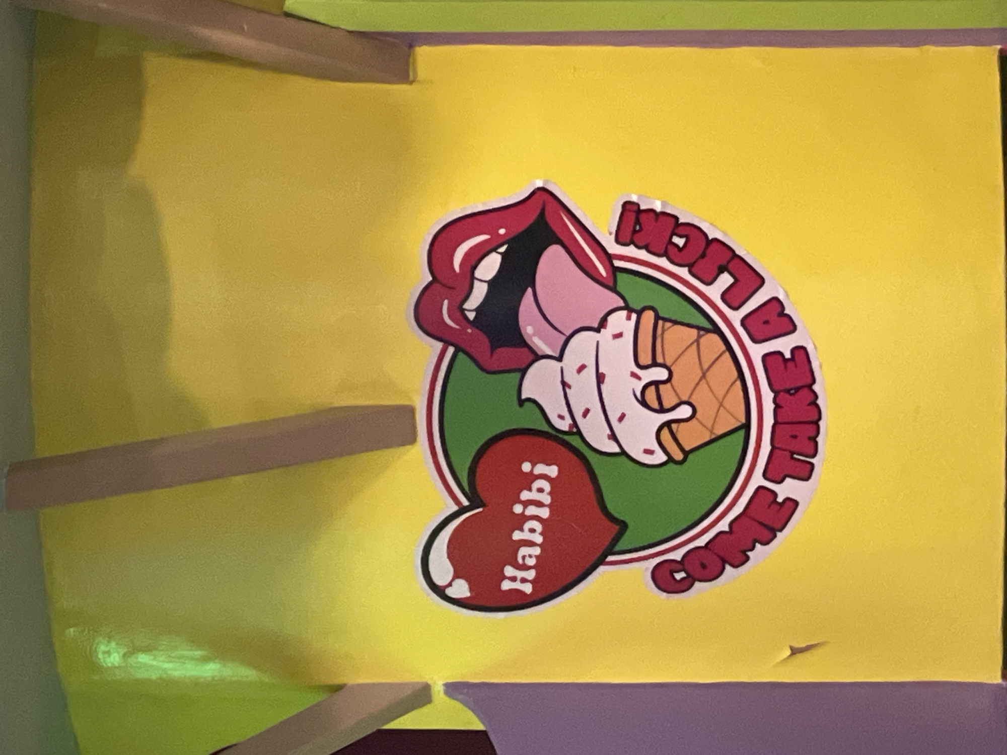 The image shows a close-up of a vibrant yellow wall decorated with a graphic sticker. The sticker features a red heart with the word "Habibi" in white text, an ice cream cone with pink frosting and sprinkles, and a pair of open red lips with a pink tongue. A circular band encircles these elements, containing the text "COME TAKE A LICK" in red lettering. The sticker is placed against a bright yellow background, with portions of a light brown wooden beam and a purple surface visible on the left and right edges of the frame, respectively. The setting is likely an outdoor mural or a decorated facade in Dumaguete, Philippines, possibly in an area known for its colorful street art or ice cream shops.