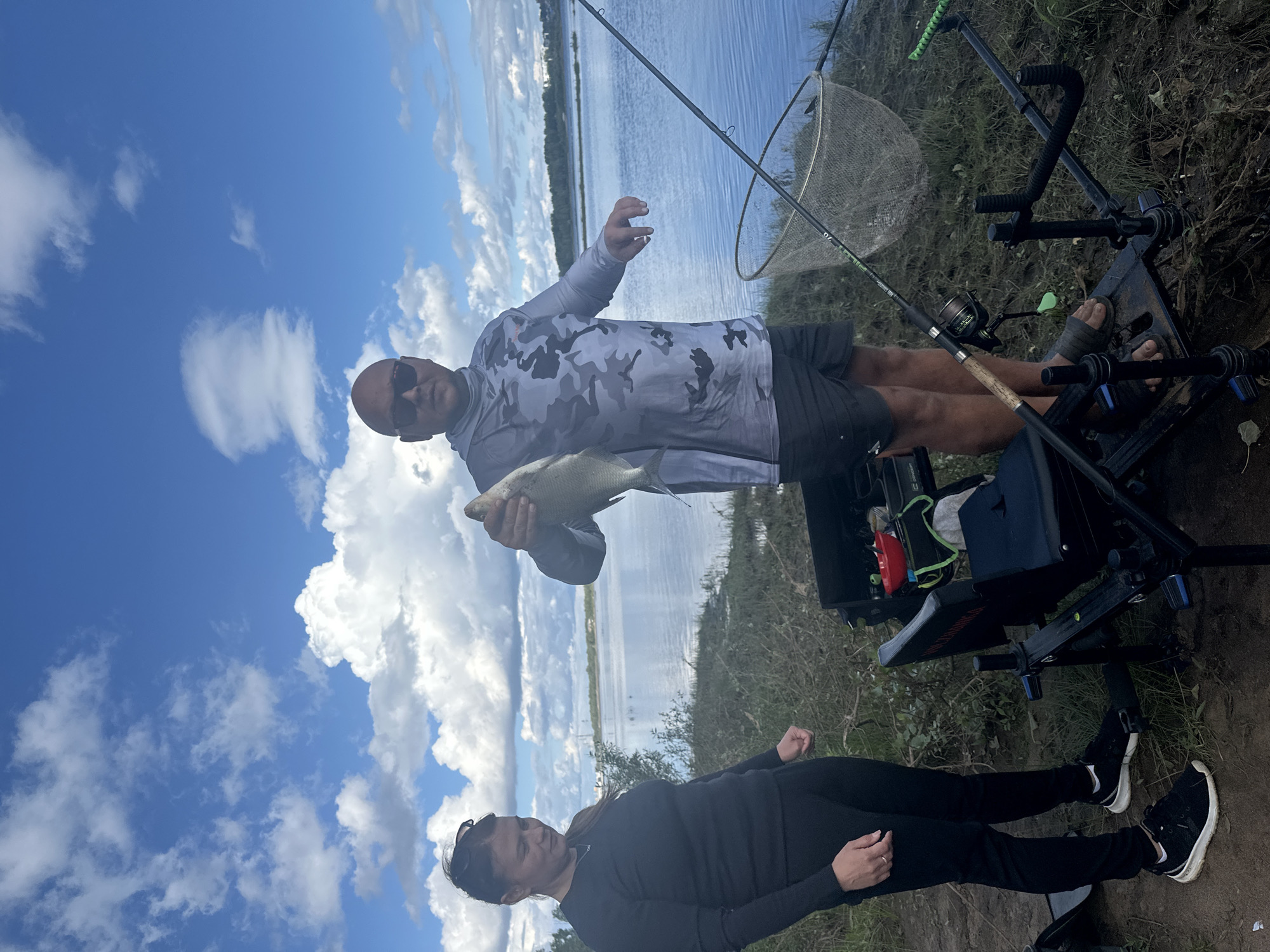 Two people are standing near a body of water in Noviy, Russia. A woman on the left is wearing a black long-sleeved shirt and black pants. A man on the right is wearing a camouflage long-sleeved shirt, black shorts, and sunglasses. He is holding a fish. In the foreground, there is fishing equipment including a fishing rod, a net, and a fishing chair. The sky is blue with white clouds, and trees line the distant bank of the water. The ground appears to be dirt and grass.