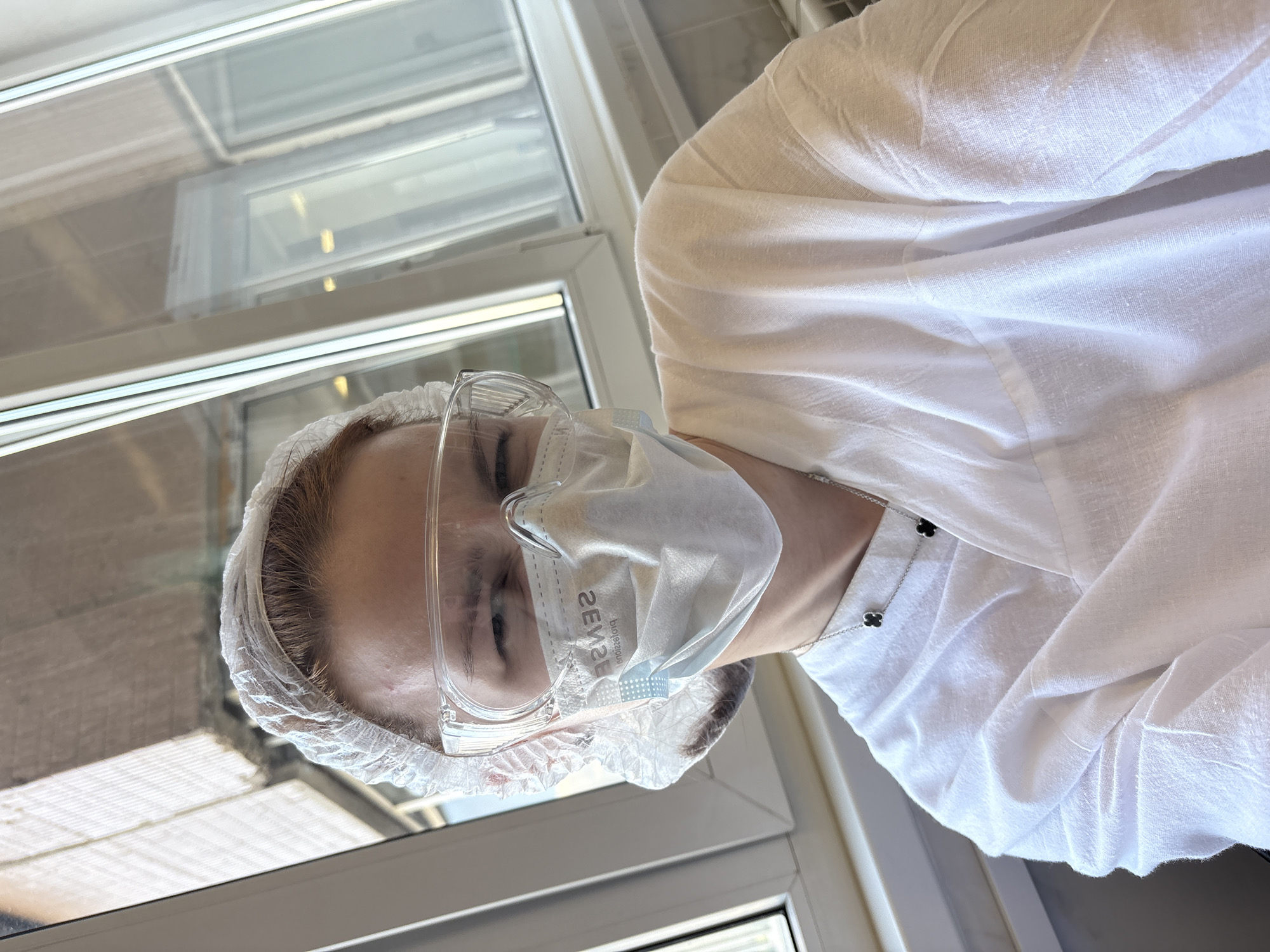 The image features an individual, likely female, wearing protective gear, captured at a tilted angle. The shot is from the chest up, focusing on the person's head and upper torso.
The person wears a transparent, disposable white hairnet, clear protective safety glasses, and a white surgical mask covering the nose and mouth. Visible text on the mask reads "SENSE biologiques", appearing reversed due to the angle. They are dressed in a loose-fitting, white long-sleeved shirt, possibly a professional uniform or lab coat, made of a textured, linen-like fabric. A silver chain necklace with two distinct black clover-shaped pendants is visible around their neck, resting against the white shirt.
The background features a large window with white frames, indicating an indoor setting. Reflections of interior structures or external architecture are visible in the window panes. The scene is brightly lit by natural light. The person's facial expression is mostly obscured by the mask and glasses, with a neutral appearance. A small blemish or red spot is visible on the forehead. No specific activity is shown beyond posing for the photograph. The attire suggests a professional or cleanroom environment, consistent with potential medical or laboratory work, possibly located in Saint Petersburg, Russia.