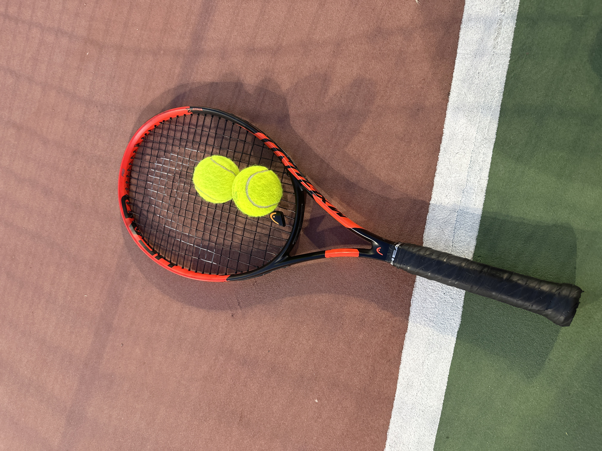 A tennis racket with two tennis balls rests on a tennis court. The racket is red and black, with the brand "HEAD" visible on the handle. The balls are bright yellow. The court surface is reddish-brown, with a white line marking the boundary of the playing area. A green section of the court is visible in the lower left corner. The lighting suggests it is daytime, with shadows cast by the racket and balls. The scene is static and devoid of any human presence. This image was taken on a tennis court in Sofia, Bulgaria.