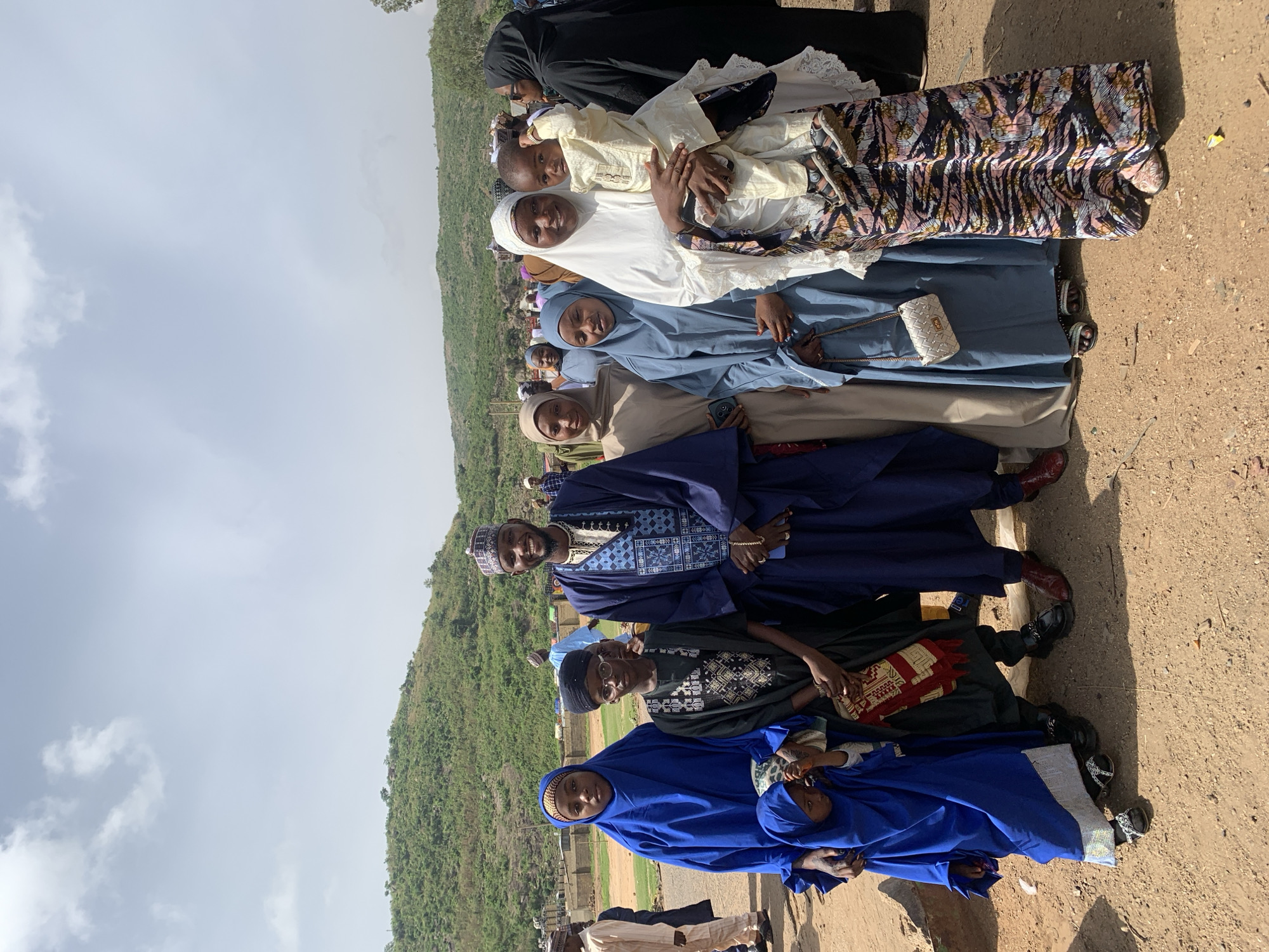 A line of people of varying ages and skin tones stand together in an outdoor setting in Naraguta Hausawa, Nigeria. The line consists of men, women, and children, who are dressed in various forms of traditional Muslim clothing. The women wear hijab and veils of different colors and patterns, including white, blue, beige, and black. The men wear traditional robes and hats, some embroidered. A young child is being held by one of the women. The background consists of a green, sloping hillside on the left and a dirt and grassy ground area. The sky is visible above, with some clouds present.