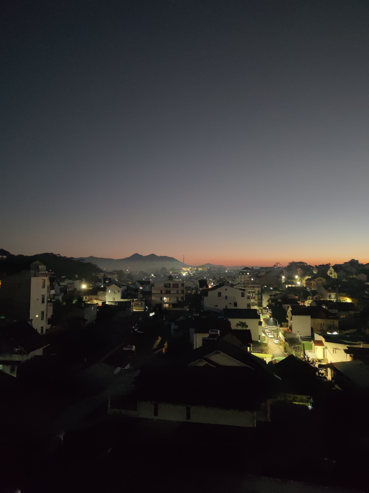 The image presents an evening view of Da Lat, Vietnam, with the city buildings spread across a hilly terrain. The sky exhibits a gradient of colors, transitioning from a dark gray at the top to a reddish-orange hue near the horizon, suggesting either sunrise or sunset. In the distance, mountains are silhouetted against the skyline. The city below is dotted with lights from buildings and streetlights, creating a network of illuminated areas amidst darker patches where the houses are in shadow. In the foreground, rooftops of houses are visible, fading into deep shadows.