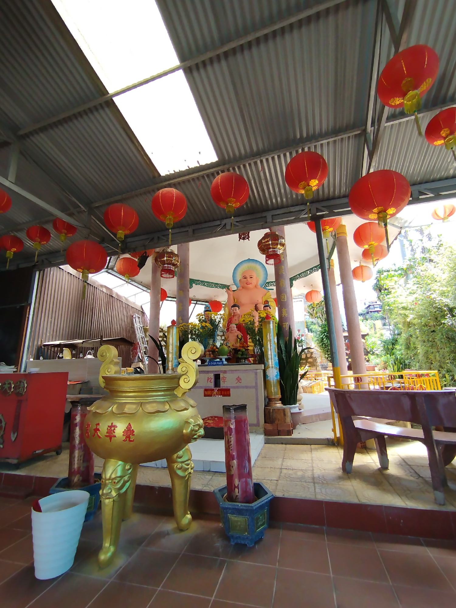 The image shows an interior view of a Buddhist shrine, likely in Da Lat, Vietnam, given the architectural style and cultural elements.
Numerous round, red lanterns are suspended from the corrugated metal roof. In the center of the scene is a raised platform, which houses a Buddha statue with a blue halo. Smaller statues and offerings are placed at its base, surrounded by flowers and greenery.
In the foreground, there is a large golden incense burner with decorative lion-paw legs. There are tall red candles in square, blue planters. A white bucket is situated near the burner. A red cabinet is partially visible on the left, and a brown wooden table with a bench occupies the right side of the scene. The floor is covered with brown square tiles. Outside the structure, there is dense vegetation.