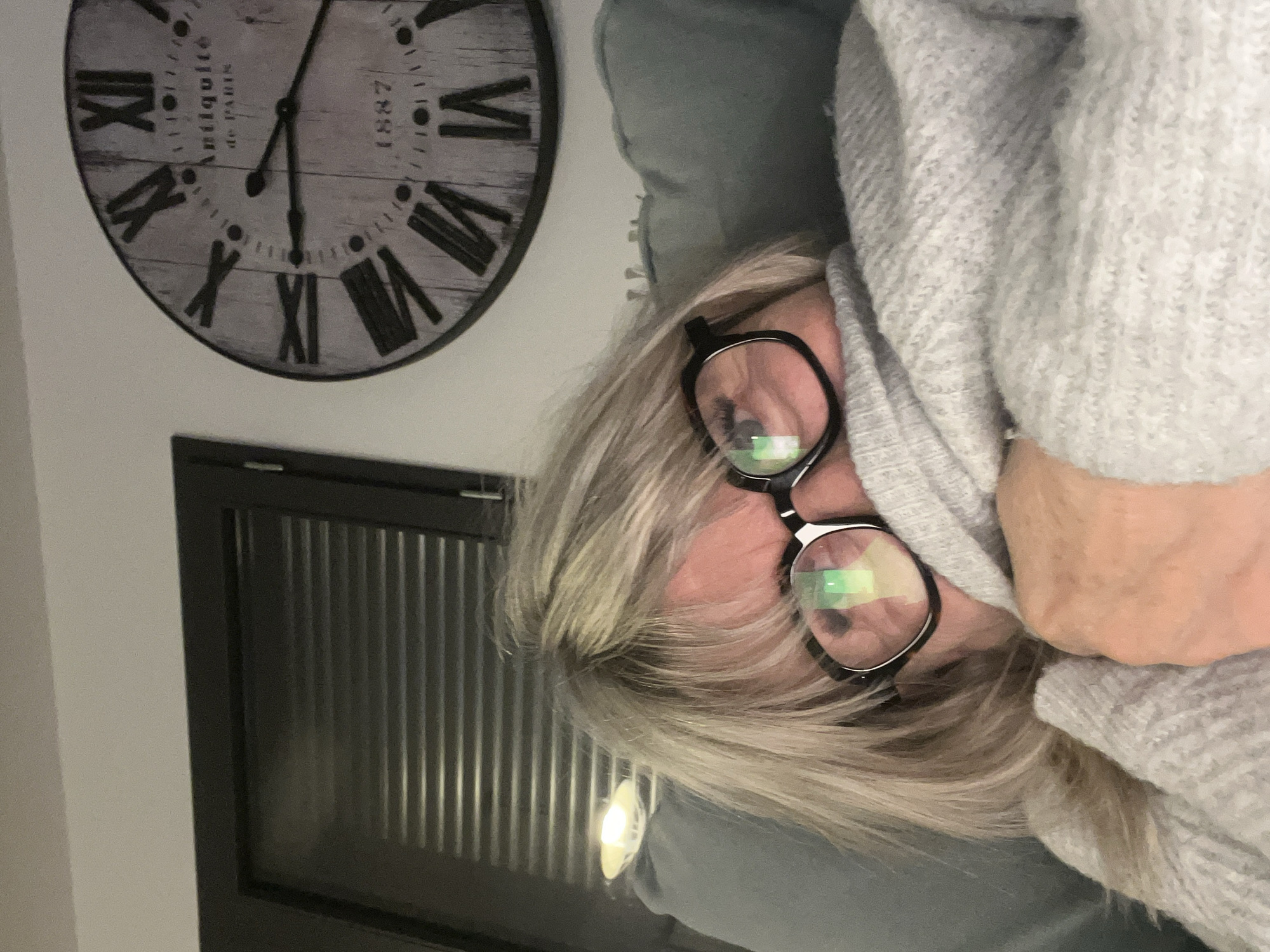 A woman is wearing glasses and a gray sweater. She appears to be lying down, possibly resting. In the background, there is a decorative wall clock with Roman numerals and a window or cabinet with vertical slats. The location in Chambœuf, France cannot be determined from the image alone.