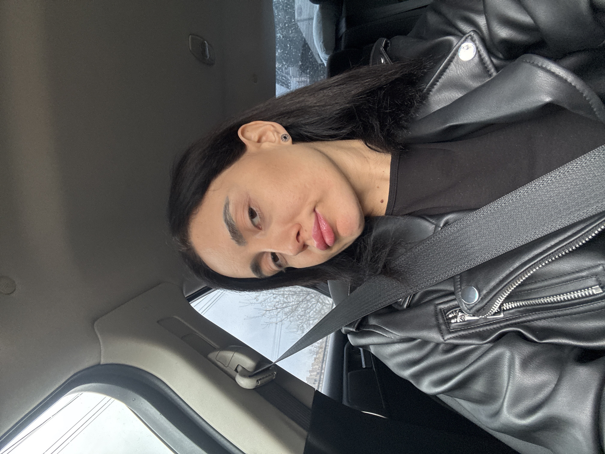 A woman is in the passenger seat of a car. She is wearing a black leather jacket and a black shirt, and has a seatbelt across her chest. She has dark hair and is looking at the camera with a slight smile. Outside the car window, it appears to be a cloudy or overcast day. Bare trees are visible outside the window. There are buildings visible in the background. The scene is potentially within Tbilisi, Georgia.