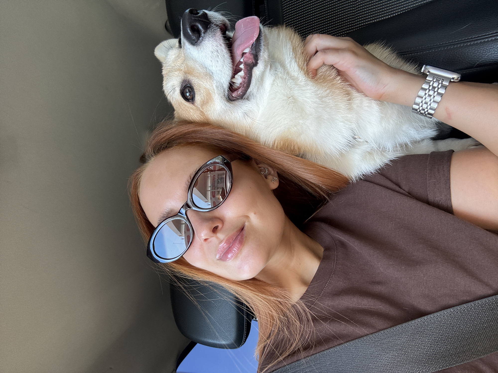 A woman in sunglasses and a brown t-shirt lies in what appears to be a car seat, with a seatbelt across her chest. Her reddish-blonde hair is spread out. A tan and white corgi dog is lying on her, with its mouth open and tongue sticking out. The woman's hand rests on the dog's neck. She is wearing a silver watch. The reflection in her sunglasses shows a building across the street with a sign in Russian, which, along with the file name, suggests that the location is the city of Chaikovsky, Russia.