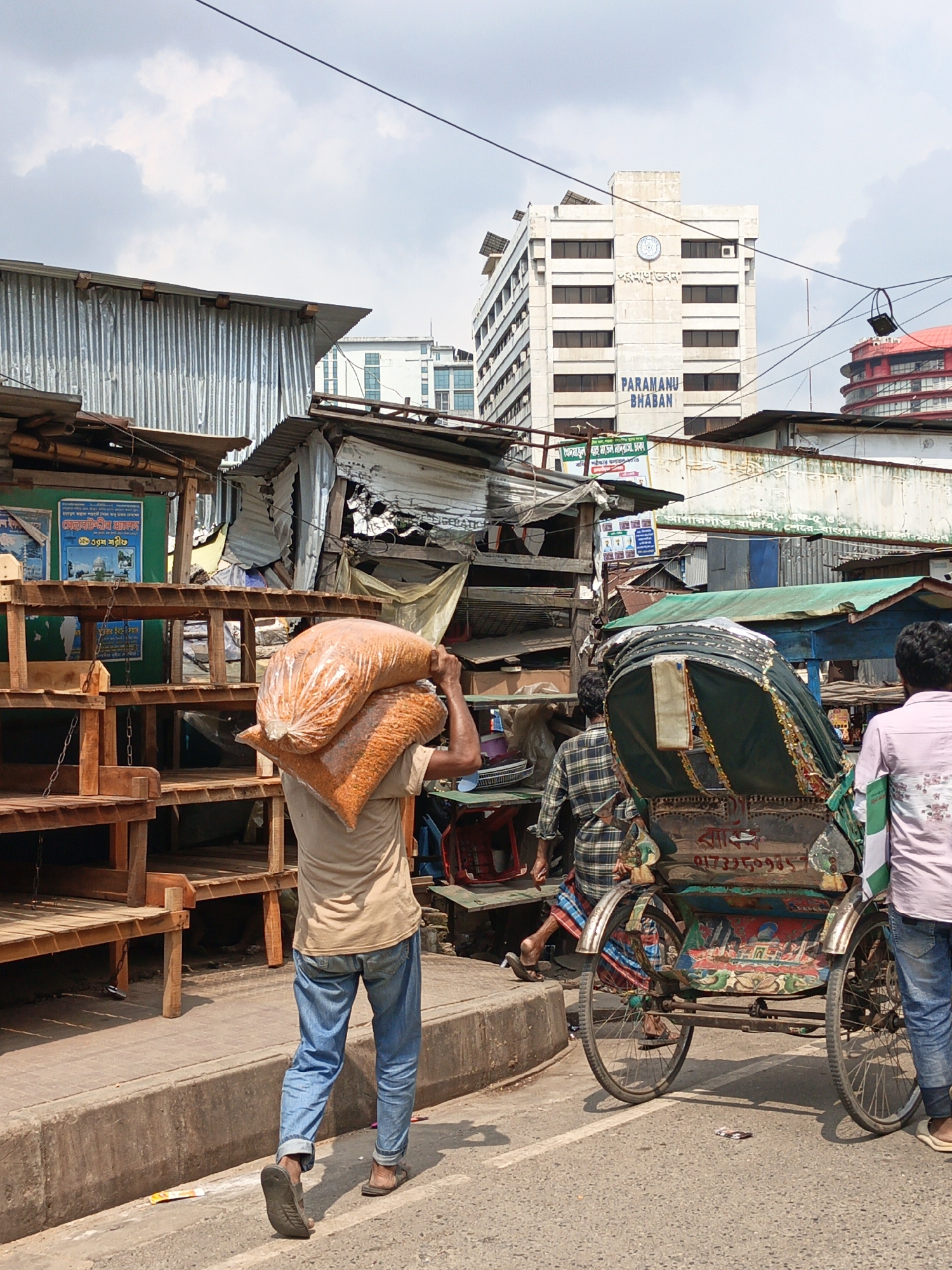 A man carries a large sack on his shoulders as he walks on a city street in Unknown, Unknown. He is wearing a light-colored shirt, blue jeans, and sandals. To his right, a colorful rickshaw is being pulled by a man in a plaid shirt and striped sarong. Another man in a pink shirt and jeans stands near the rickshaw.
In the background, there are several buildings, including a modern white building with the words "PARAMANU BHABAN" on it. There are also some makeshift structures and stalls along the side of the road. The sky is cloudy.