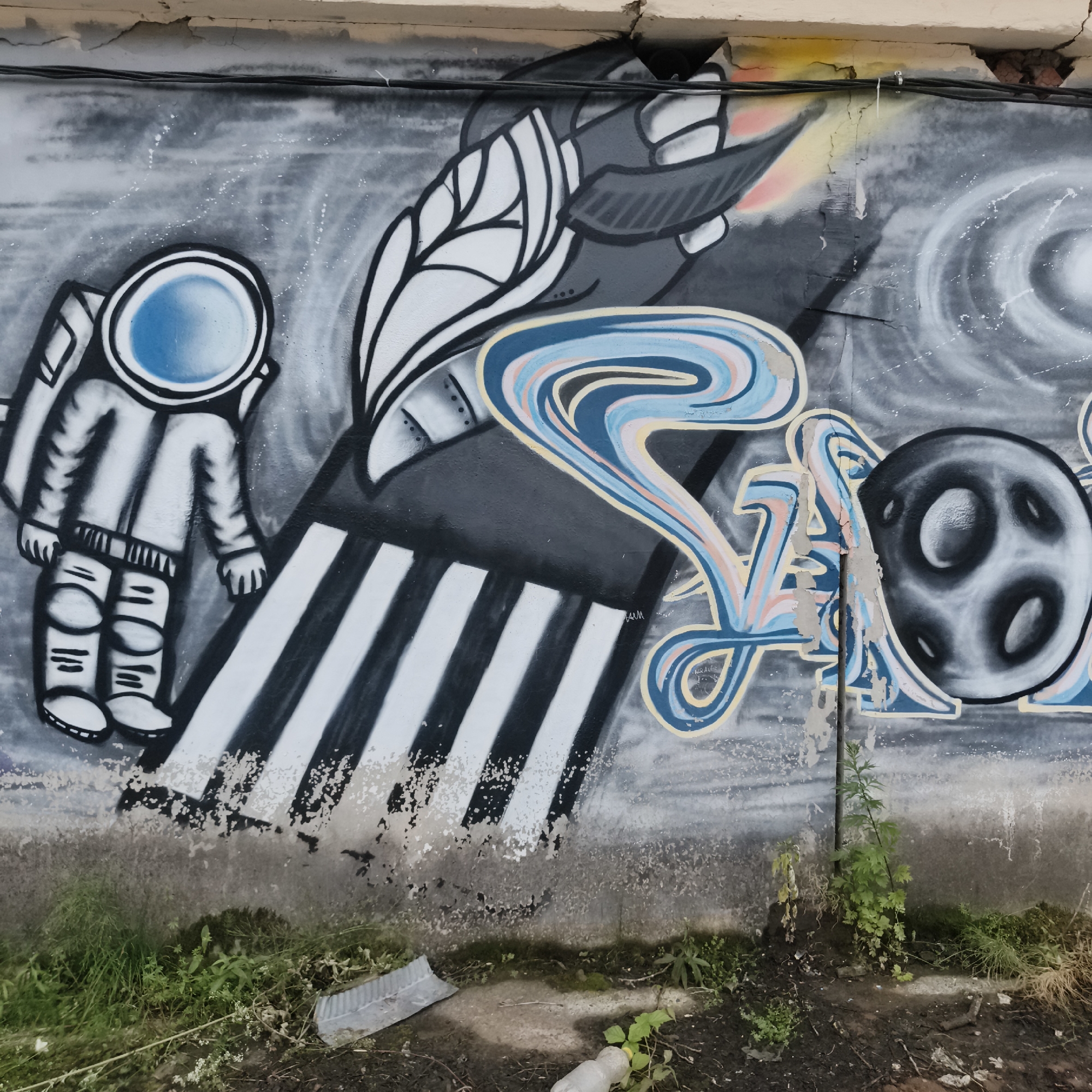 This media file depicts a graffiti mural on an outdoor wall. The mural's central subject is an astronaut in a white spacesuit with a blue visor, standing on a striped surface that resembles a runway or stepping stones. To the right of the astronaut, a stylized rocket is depicted with white leaf-like elements and dark gray body, leaving a trail of colorful smoke or flames in shades of yellow, pink, and orange. The background features swirling gray and white patterns, suggestive of outer space or cosmic phenomena.
Further to the right, abstract, flowing shapes in blue, pink, and yellow are painted, possibly representing cosmic energy or stylized text. A dark gray sphere with circular indentations, resembling a planet or moon, is positioned to the far right of the abstract shapes. The graffiti is painted on a weathered concrete wall with visible cracks and peeling paint.
The scene is set outdoors, with vegetation, including grass and small plants, growing at the base of the wall. A discarded plastic bottle is visible in the foreground. The lighting suggests daytime, with no specific indication of weather conditions. The location context is identified as Chaikovsky, Russia. There are no clearly legible words or text visible within the graffiti, apart from a small, illegible tag near the abstract shapes. The overall theme of the artwork appears to be space exploration and cosmic adventure.