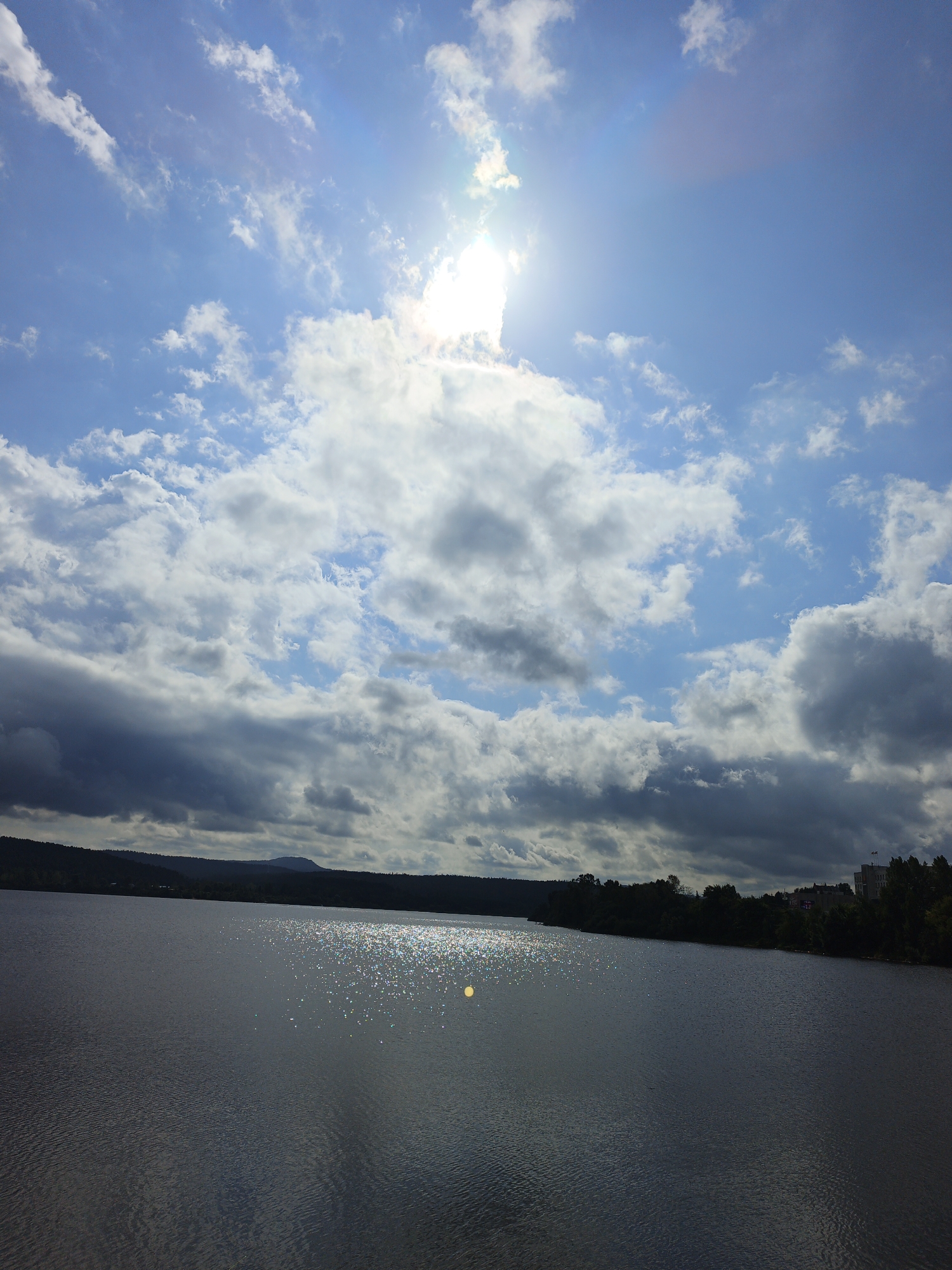 The image captures a wide view of a lake under a dynamic sky with bright sunlight and scattered clouds. The sun is high and partially obscured by clouds, creating a luminous effect and casting reflections on the water's surface. The water is calm with subtle ripples. A dark, tree-lined shoreline forms the backdrop, with a distant, hazy mountain range. On the right side of the image, a few buildings and more trees are visible along the shore, suggesting a settlement near the water. The scene appears to be a natural landscape within or near a city. This description is consistent with a view of the Zlatoust reservoir, also known as the Zlatoust Pond, located in the city of Zlatoust, Russia.