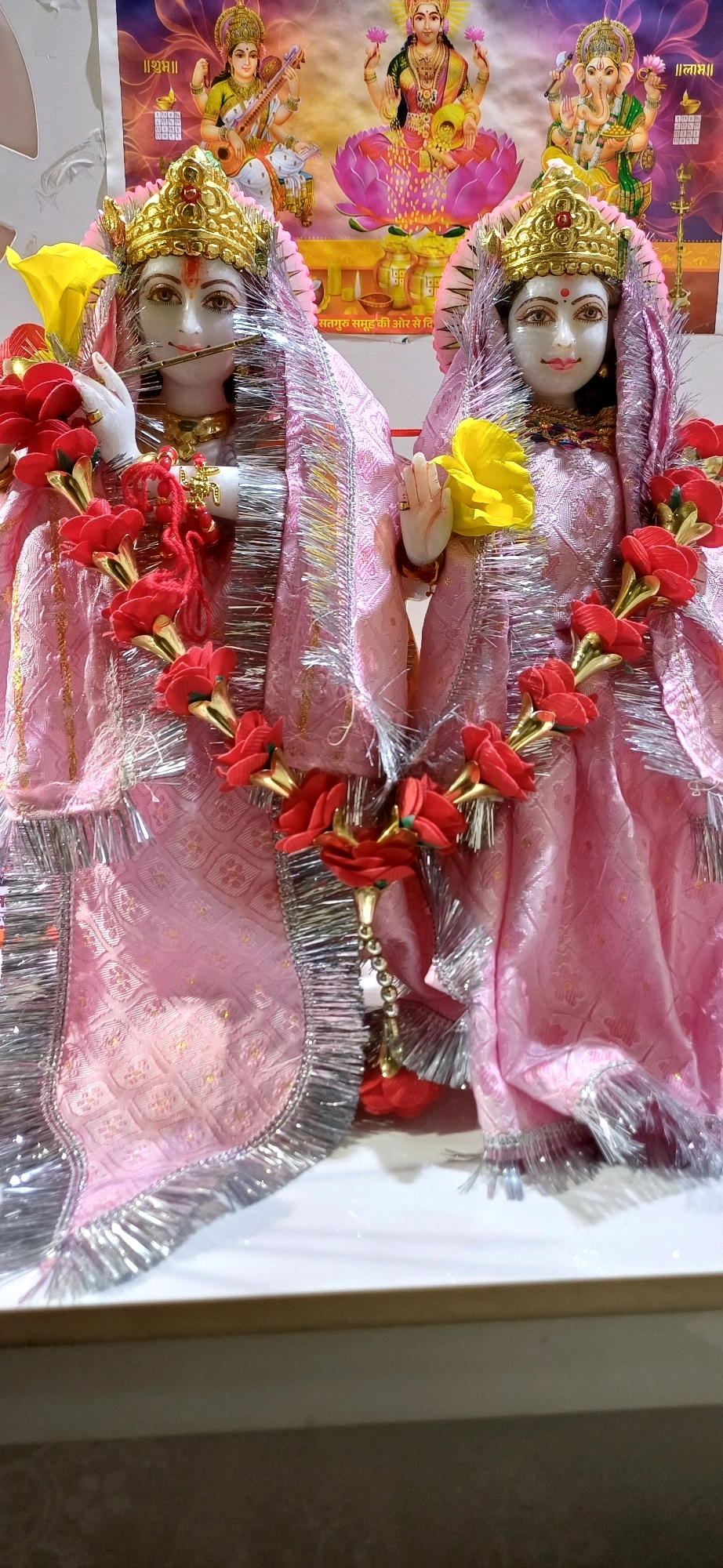 The image displays two ornate statues of Hindu deities, likely Radha and Krishna, adorned in pink silk garments with silver fringe trim. Both statues wear golden crowns. The statue on the left, assumed to be Krishna, holds a flute to its lips, while the statue on the right, assumed to be Radha, has its hands in a gesture of blessing or offering. Red and gold floral garlands are draped around the necks and bodies of the statues. In the background, a religious poster depicts several Hindu goddesses, including Lakshmi and Saraswati. The statues are placed on a white surface which appears to be a shelf or altar. The lighting suggests an indoor setting. The photograph was taken in Ajmer, India, inside a religious establishment, possibly a temple or a home altar dedicated to Hindu worship.