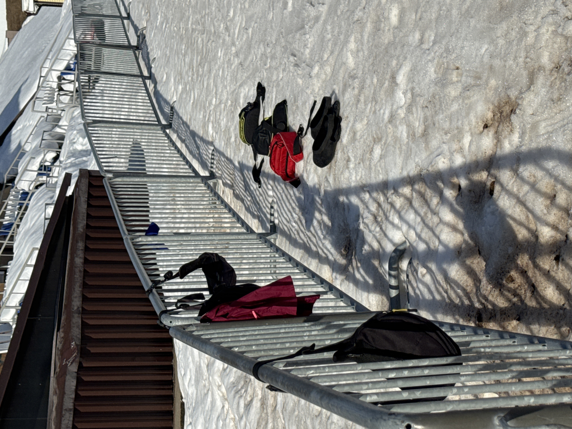 An outdoor scene in Прикамский, Russia, depicts a snow-covered area under clear skies. A line of galvanized metal temporary barriers extends diagonally from the lower left to the upper right. Hanging on the barriers are a black bag with a yellow zipper pull, a burgundy-red jacket, and a partially visible second black bag. On the patchy, dirty snow directly adjacent to the barriers, four items are laid out: a black bag with yellow accents, a red bag, another black bag, and a pair of black, flat-soled shoes or slippers. Long shadows from the barriers are cast onto the snow. In the background, to the left, is a brown corrugated metal wall, and to the right, snow-covered spectator seating or bleachers with some exposed blue seats are visible. No individuals are present in the image.