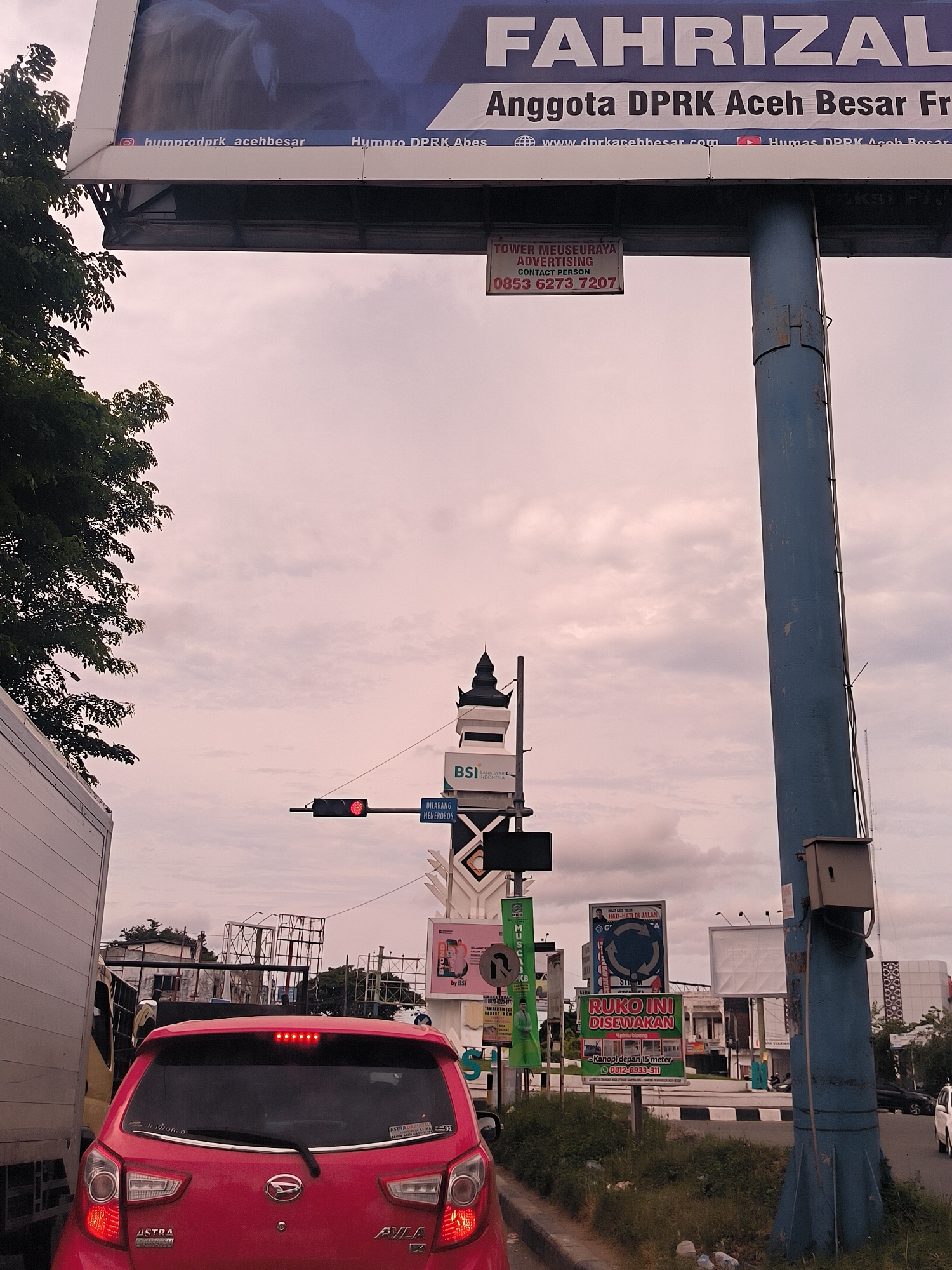 The image presents an urban street scene in Lambaro, Indonesia, from the perspective of a vehicle stopped in traffic. In the foreground, the rear of a red Daihatsu Ayla car is prominently featured, with its brake lights illuminated. To the left, part of a white truck or van is visible, suggesting traffic congestion.
Overhead, a large billboard displays the name "FAHRIZAL" and identifies him as an "Anggota DPRK Aceh Besar," accompanied by social media handles such as "humprodprk_acehbesar" and a website "www.dprkacehbesar.com." Directly below this billboard, a smaller sign advertises "TOWER MEUSEURAYA ADVERTISING" with a contact person and phone number "0853 6273 7207."
In the mid-ground, a red traffic light is visible, confirming the stop in traffic. Below it, a road sign clearly states "DILARANG MENEROBOS" (Do Not Break Through/Do Not Enter). A decorative structure, possibly resembling a minaret or traditional turret, stands near a sign for "BSI Bank Syariah Indonesia." The sky is overcast, providing soft, diffused lighting, indicative of a cloudy day or a specific time like late afternoon.
The background reveals several other commercial billboards and signs along the roadside. Notable among them is one offering "RUKO INI DISEWAKAN" (This shop for rent) with details like "Kanopi depan 15 meter" and a contact number "0812-6933-311." Another sign encourages caution with the phrase "HATI-HATI DI JALAN" (Be careful on the road). The scene depicts a typical Indonesian street environment filled with advertising and traffic infrastructure, momentarily at a standstill.