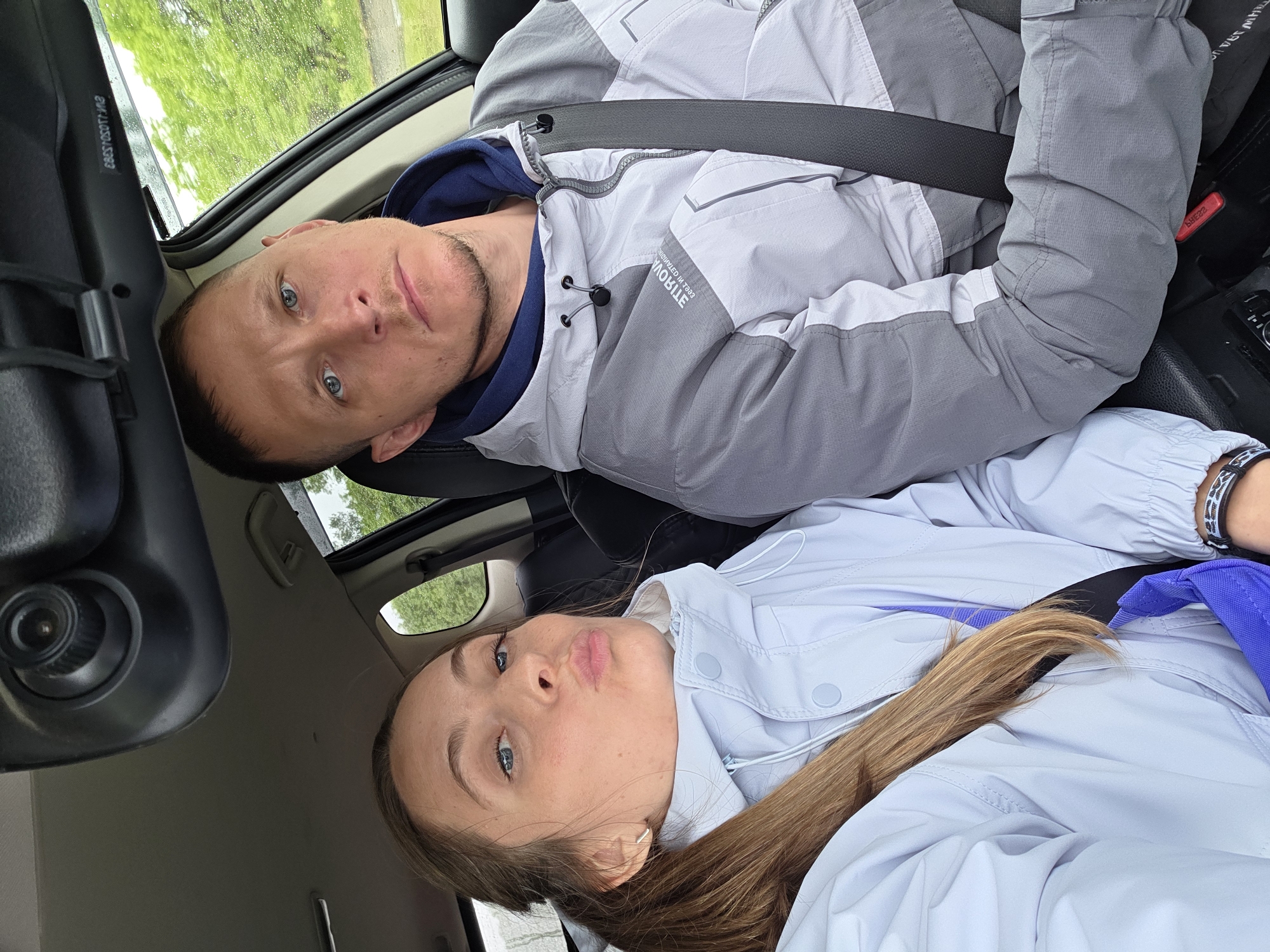 Two individuals, a man and a woman, are seated in the front seats of a car, seen from an upward angle and rotated counter-clockwise. They both appear to be wearing seatbelts.
The man, on the left, has short dark hair, blue eyes, and light stubble on his face. He is wearing a grey and white athletic jacket over a dark blue hoodie. Text on his jacket, which is mirrored, shows "MOBILE" and possibly "TERAVOVA" or similar letters above it. He looks directly at the camera with a neutral expression. Near his head, mirrored text, possibly a VIN or serial number "EB037SSOTTL113", is visible on the car's interior trim.
The woman, on the right, has long brown hair and blue eyes. She is wearing a light blue or lavender jacket and is making a "pouty" or "duck face" expression towards the camera. A black and white patterned bracelet is visible on her wrist, and a blue strap (possibly from a bag) is seen near her.
The interior of the car features dark and light-colored components. Through the car windows, lush green foliage and trees are visible, suggesting an outdoor, natural setting. The reflections on the window and diffused light indicate overcast weather, possibly with some wetness or condensation. An overhead console with a control knob is visible in the car's ceiling. A red seatbelt release button is visible on the passenger side.
The image captures a casual moment of two people in a car, likely taken as a selfie or by someone in the back seat, in Бабстово, Russia.