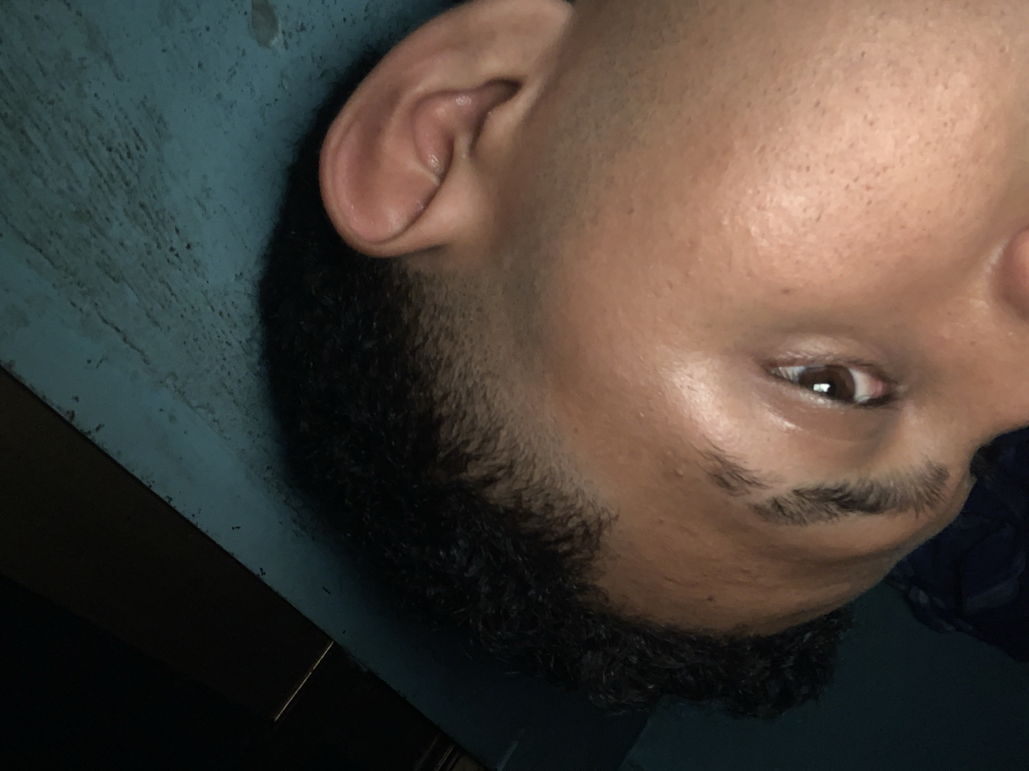 A close-up portrait captures the right side of a person's face, rotated so the head appears sideways or inverted. The subject has medium brown skin, a dark brown eye that looks directly into the camera, and a thick, dark eyebrow. Dark, curly hair is visible along the hairline and sideburn area, styled with a noticeable fade. The ear is prominent and detailed. The skin texture, including visible pores on the forehead and cheek, is apparent. The background features a textured, dark teal or blueish-green surface, suggesting an indoor setting, possibly a wall or furniture. Darker, linear elements are present in the lower left portion of the image, further hinting at an interior environment. The lighting is somewhat dim, casting subtle highlights on the skin. The person is posing for the photograph. This scene takes place in Santa Ana, Venezuela. No text is visible.