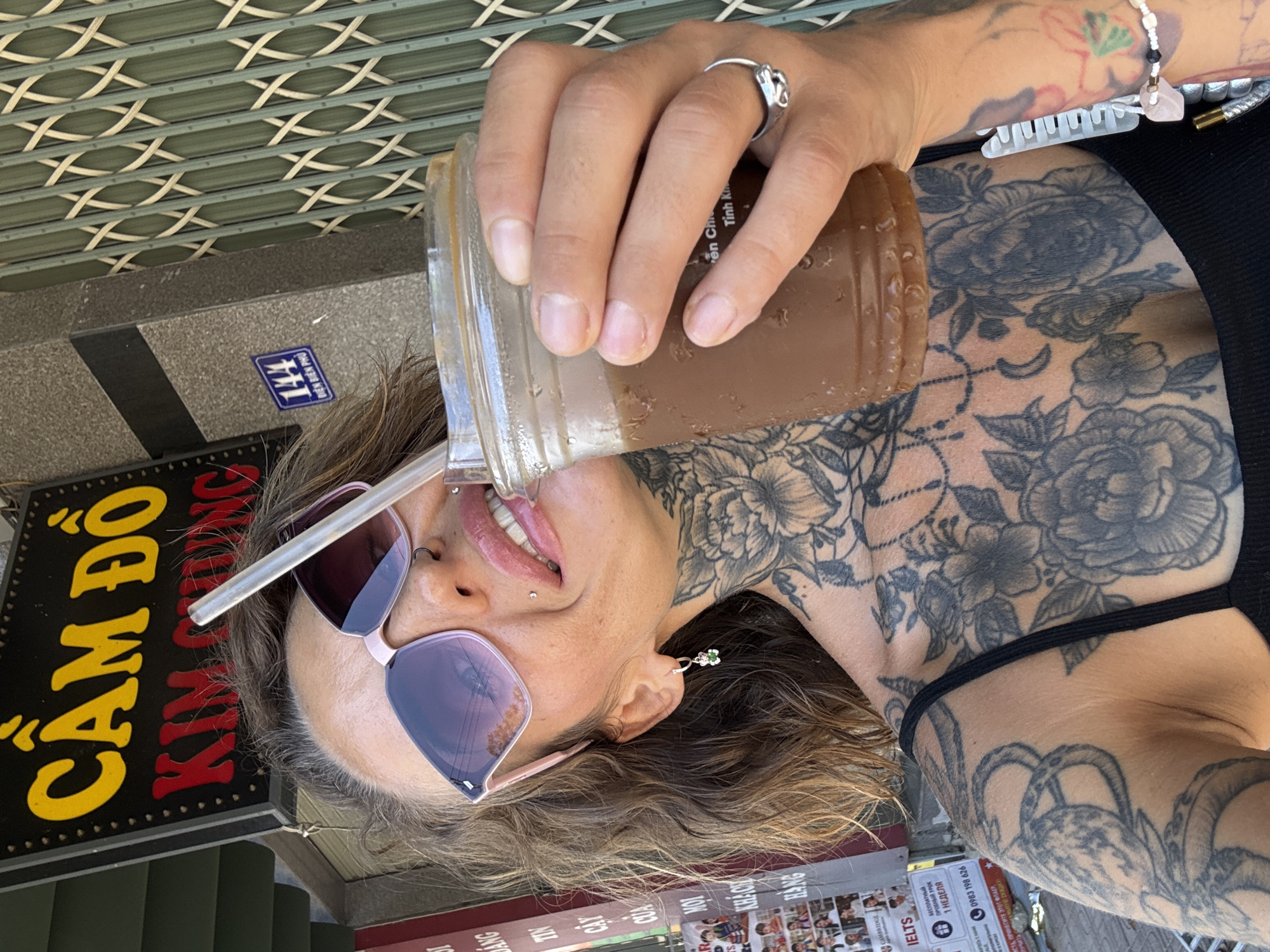 The image shows a woman with extensive tattoos on her neck and chest, holding a plastic cup of iced coffee. She is wearing sunglasses and a black tank top. The background suggests an outdoor setting in a city, with a sign in Vietnamese characters, a building with a metal grate, and a poster board with multiple images. The sign in the background reads "CẨM ĐÔ KIM CHUNG", which appears to be a business name, likely a jewelry store or similar establishment, given the Vietnamese word "CẨM" (gem/jade) and "KIM" (gold). The number "144" is visible on a blue plaque on the wall, possibly indicating a street number. The overall scene is a candid portrait of the woman enjoying a drink outdoors in a Vietnamese urban environment.