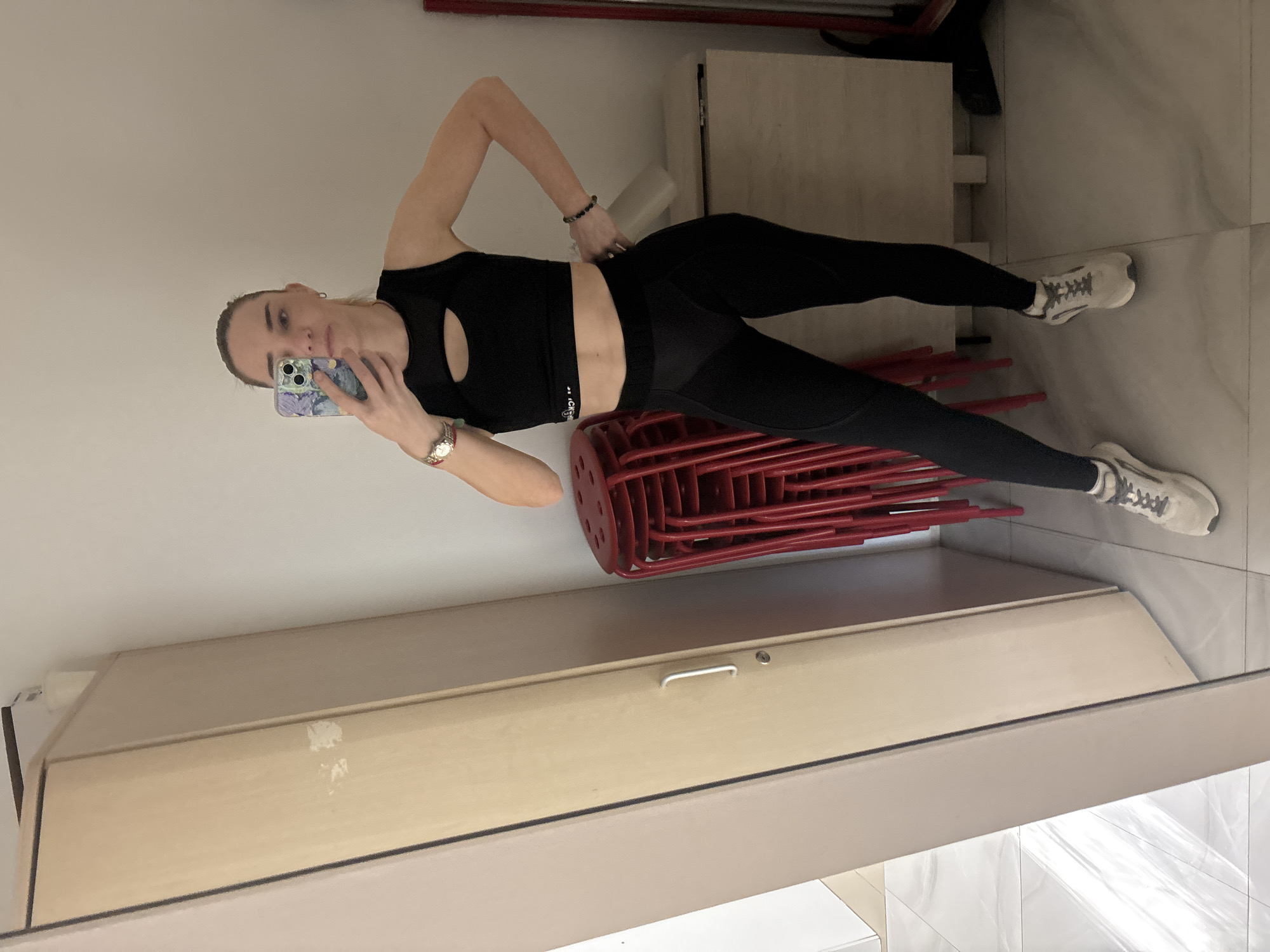 A woman in black athletic wear, including a crop top and leggings, stands in front of a mirror and takes a selfie. She is wearing light-colored athletic shoes and has a watch on her left wrist and bracelets on her right. Behind her, a stack of red stools is visible, as is a wooden closet or wardrobe. The room appears to be a residential or gym space. The overall impression is that of a casual, personal moment captured for social media.