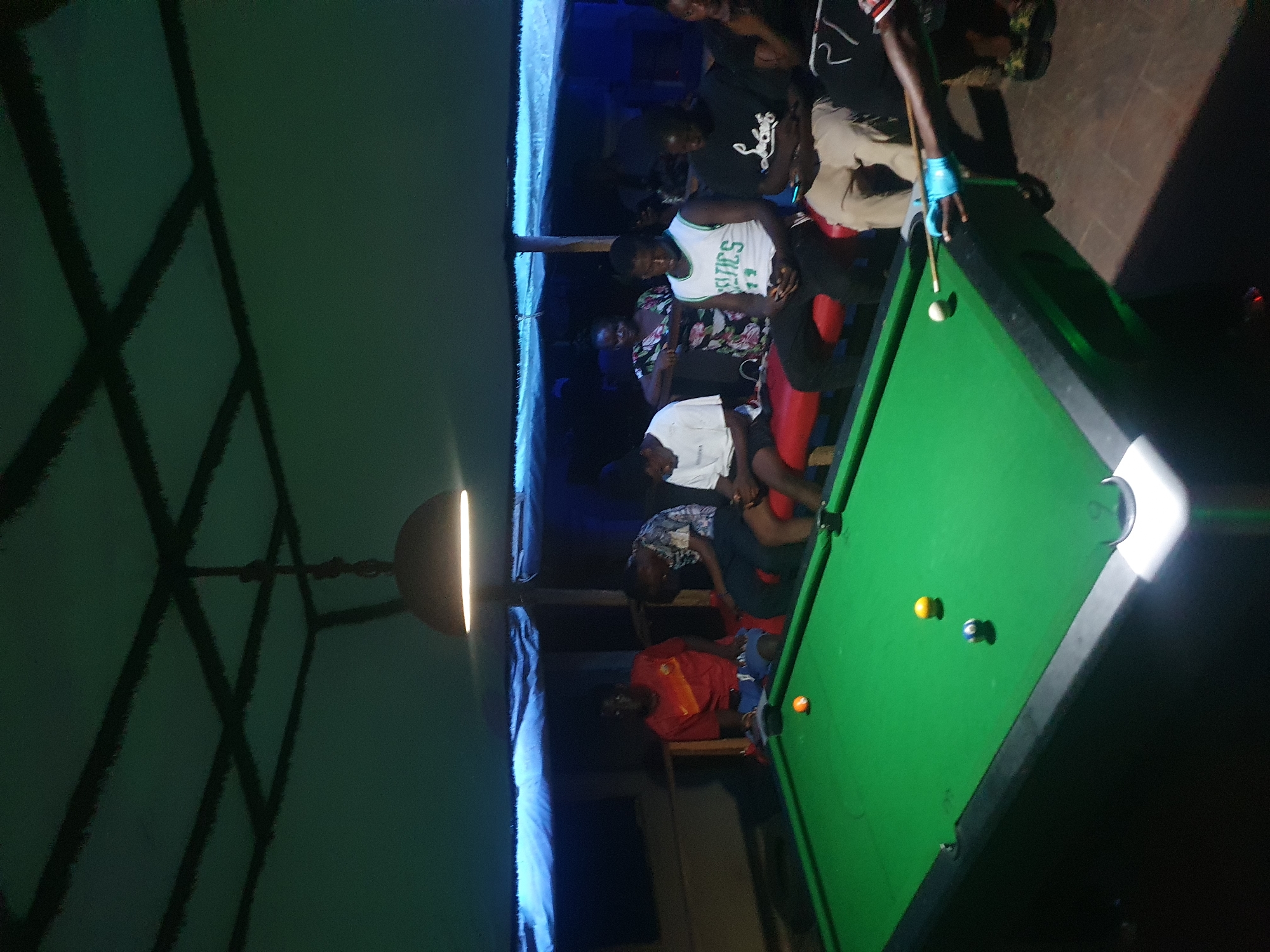 This media file captures a lively indoor scene, likely in a bar or recreational hall in Lokoja, Nigeria, during the evening or night. The image is oriented vertically, showing a dimly lit space where several individuals, predominantly men, are gathered around a pool table.
The central activity revolves around a game of pool. One person, wearing a light blue glove on their left hand, is actively taking a shot on a green pool table. The table has a white cue ball, an orange ball, a yellow ball, and a blue striped ball visible. Other people are standing or seated around the table, observing the game, contributing to a social atmosphere.
The setting features dark walls and a ceiling with a prominent grid or lattice pattern. A circular pendulum light fixture hangs from the ceiling, casting a bright, direct light downwards, illuminating parts of the room and creating reflections on the shiny elements of the pool table. The overall lighting is artificial and dim, enhancing the evening ambiance.
Visible details include:
* Several individuals of varying ages, dressed in casual attire.
* One person wears a white basketball jersey with "LTICS" visible on the front, likely "CELTICS".
* Another person wears a red shirt.
* Seating, possibly benches or low chairs, is present near the pool table.
* The floor appears to be a dark, solid surface.
The image conveys a sense of casual social interaction and entertainment centered around the game of pool.