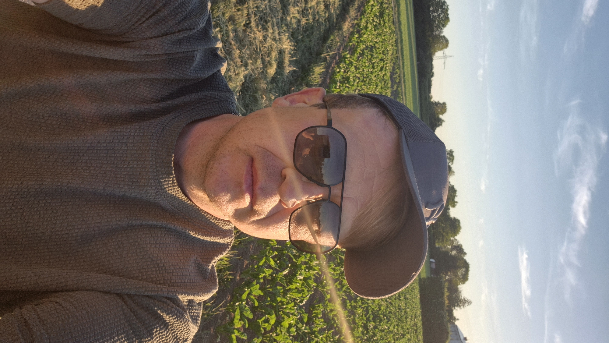 A man wearing sunglasses and a baseball cap is standing in a field in Ergolding, Germany. He is wearing a brown waffle-patterned long-sleeved shirt. The field appears to contain both freshly cut grass and green vegetation. The sky is light blue with some thin clouds. In the background, trees, power lines, and buildings are visible.