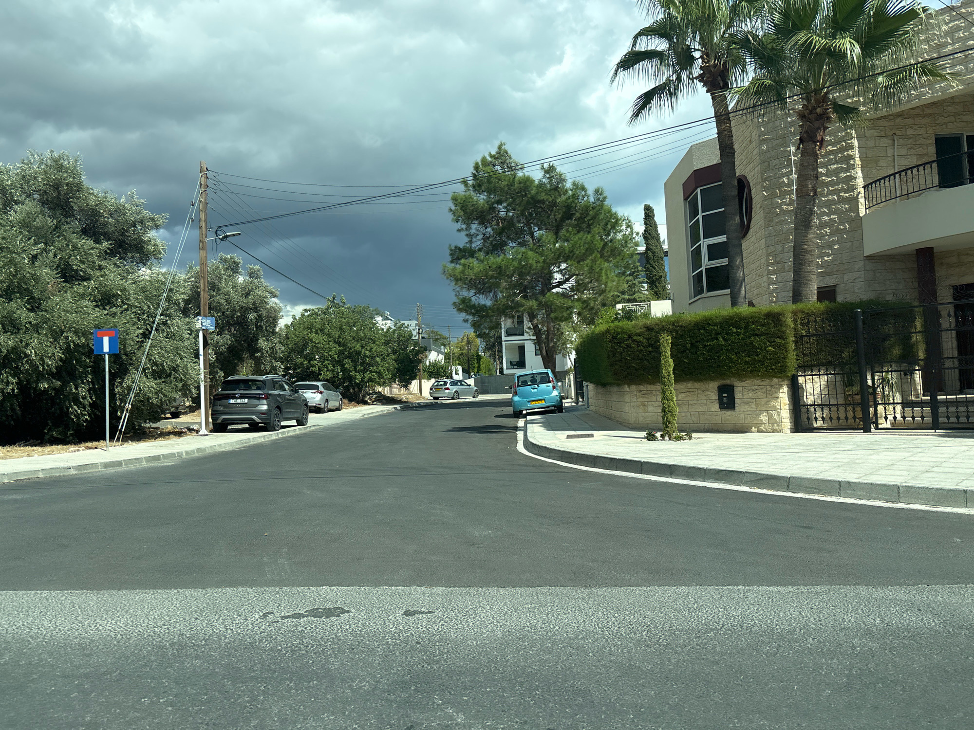 The image shows a residential street in Germasogeia Municipality, Cyprus, on a cloudy day. A grey Volkswagen T-Roc is parked on the right side of the street, followed by a silver Volkswagen Golf and a light blue car. A single palm tree and a manicured hedge are visible in front of a modern beige stone house with a black metal fence. Telephone poles and power lines run along the street. A blue and red sign indicating a dead end is posted on a pole to the left, next to a dense green tree. The road is paved and appears to be in good condition, with a pedestrian crossing visible in the foreground. The sky is overcast with dark clouds, suggesting impending rain.