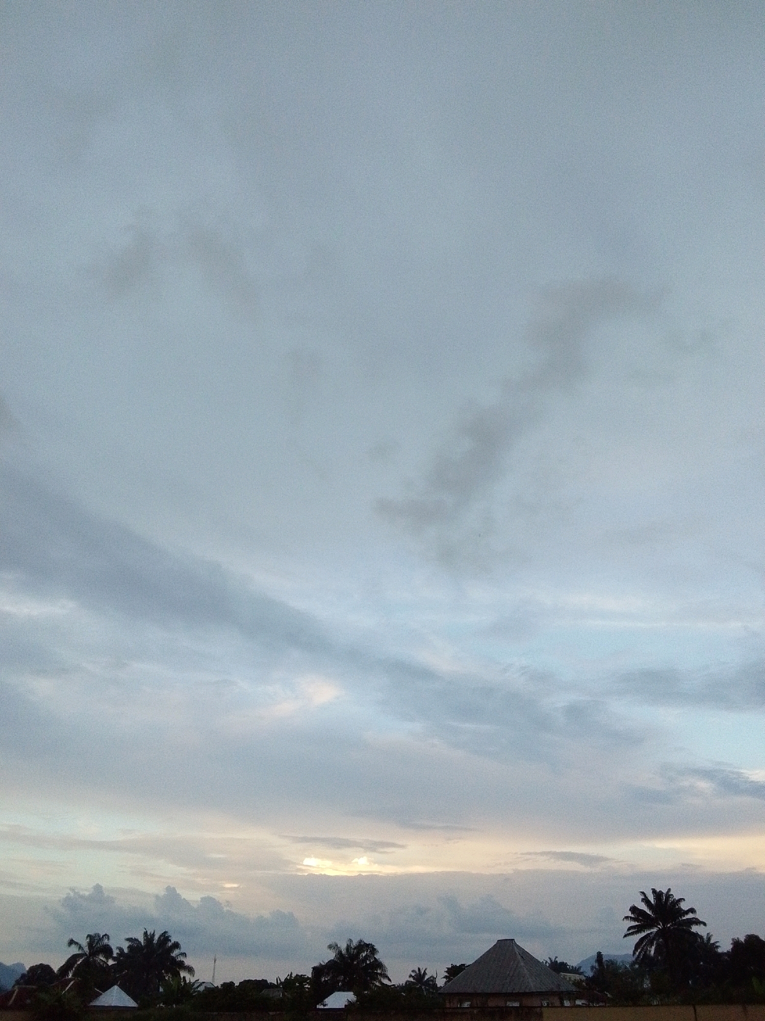 The image depicts a cloudy sky over a rural landscape in Serti, Nigeria. The sky is filled with a mixture of light gray and white clouds, with a hint of pale yellow near the horizon where the sun is obscured. In the foreground, silhouetted against the sky, are various trees, including palm trees, and several structures. One prominent structure is a building with a conical, thatched roof. The overall impression is a serene, natural environment with scattered signs of human habitation.