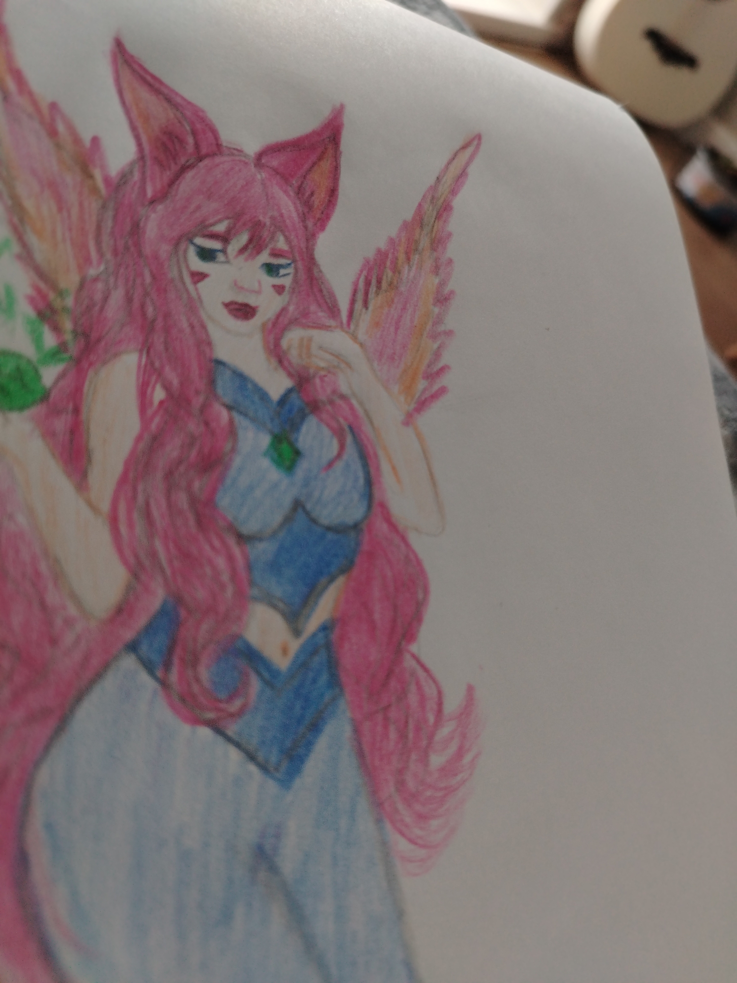 A drawing of a pink-haired anime-style character with fox ears and multiple tails is on a white piece of paper. The character is wearing a blue dress or top with a green jewel. The drawing is partially obscured by a white object in the upper right corner, possibly a roll of tape. The background is out of focus and appears to be a neutral-toned surface. There is no indication of a location in Izhevsk, Russia.