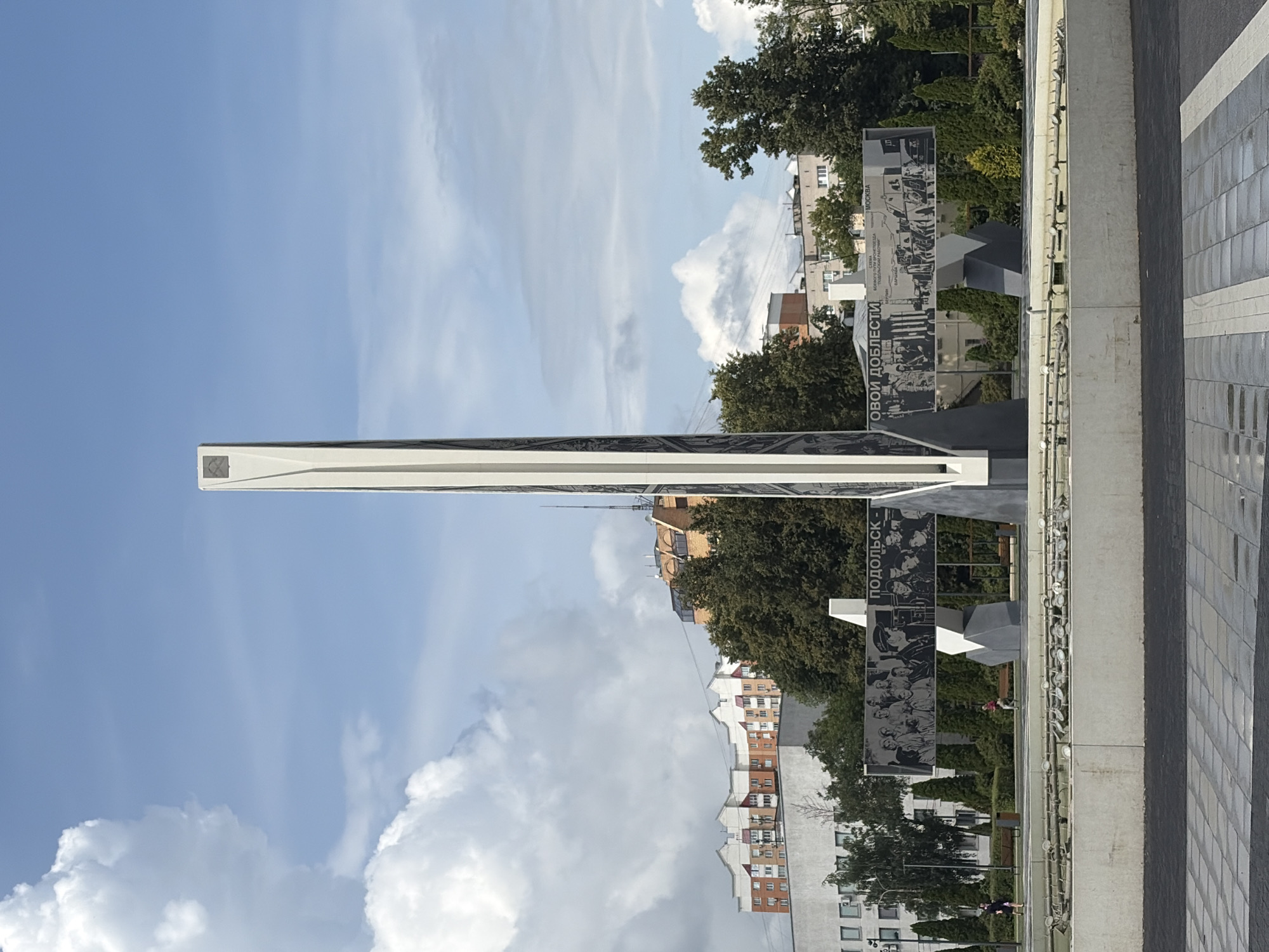 The image shows a monument in the city of Podolsk, Russia, dedicated to the Battle of Moscow. The monument is a tall, white, obelisk-shaped structure with dark accents, positioned in the center of a plaza with a fountain in the foreground. A large black and white mural depicting scenes from the battle is mounted on a structure behind the obelisk, with text in Russian visible. The plaza is paved with stone tiles and surrounded by green trees and bushes. In the background, there are several residential buildings and some taller structures, indicative of an urban environment. The sky is blue with scattered white clouds. A few people are visible in the plaza, walking or standing, and some benches are present.