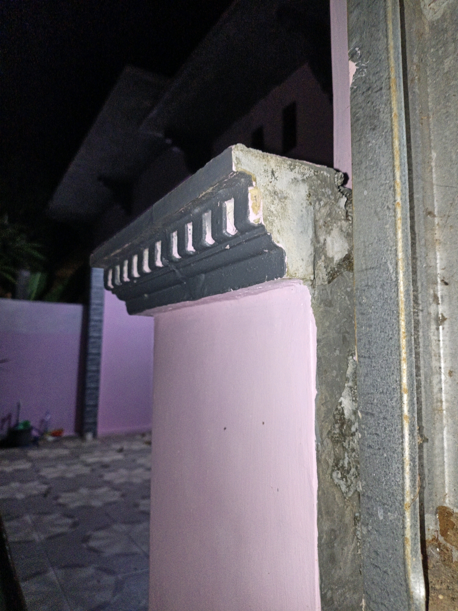 The image shows a close-up view of a gate post, likely in Cikal, Indonesia. The post is made of concrete and is partially covered by a corrugated metal structure. The concrete has a decorative molding at the top, painted dark gray with white accents. The pink wall of a building is visible behind the post, and a paved walkway with a patterned design is in the background. The lighting suggests it is nighttime or a very dark, overcast day. There are no people or animals visible in the image. The scene appears to be an outdoor area adjacent to a residential property.