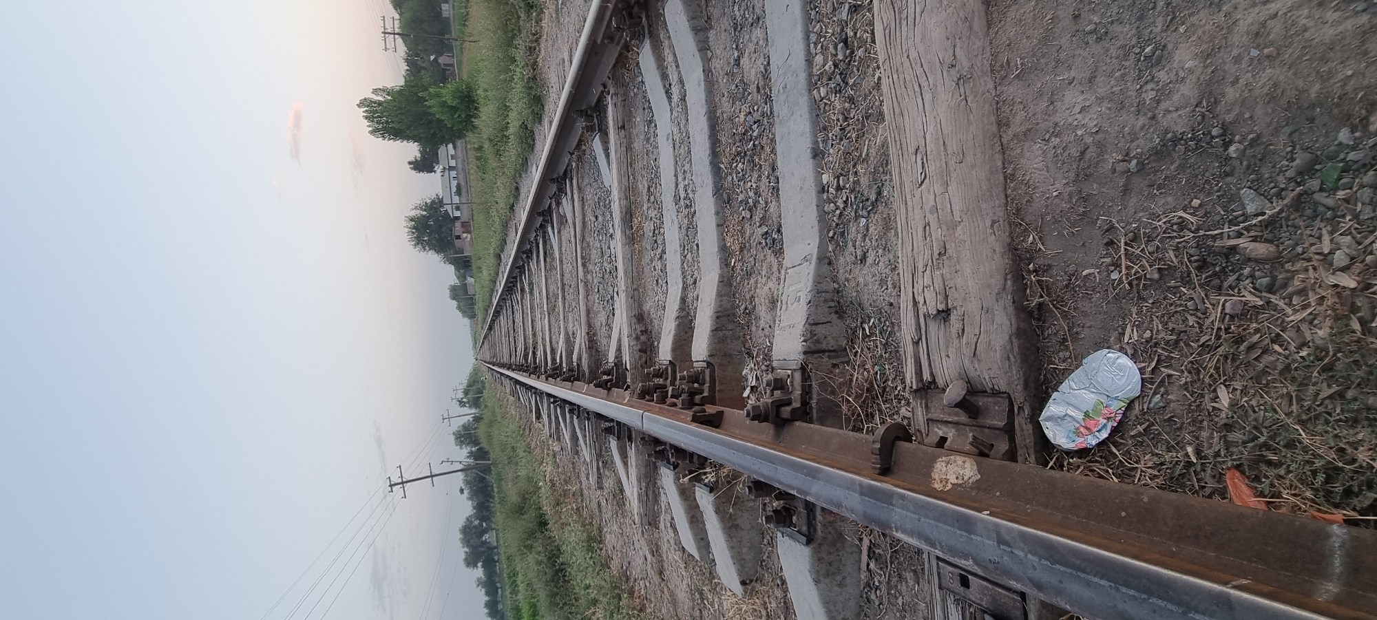 The image shows a railroad track stretching into the distance. The track consists of two steel rails supported by concrete and wooden sleepers. Electrical poles with wires are visible along the side of the tracks, and there is vegetation and some buildings in the background, suggesting a rural or semi-urban environment. A crumpled metallic object, possibly a can, with a floral pattern is lying on the ground near the tracks. The sky is clear with a slight haze, and the lighting indicates it might be late afternoon or early morning. There are no people visible in the image. The scene is located near the city of Khovaskor, Tajikistan.