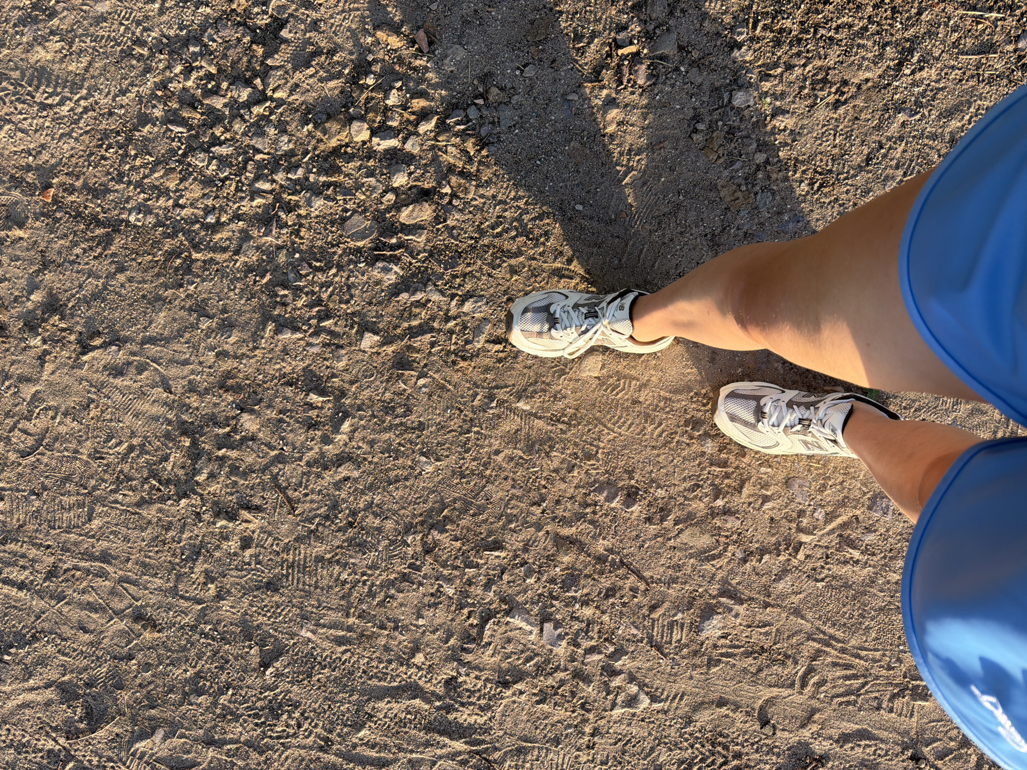 An aerial perspective captures a person's legs and feet walking on a dusty, gravel path in Ulbroka, Latvia. The ground is textured with numerous footprints, indicating recent activity. The subject is wearing white and grey athletic shoes and blue athletic shorts. The lighting suggests it is either early morning or late afternoon, casting distinct shadows on the path. The environment appears natural and unpaved, characteristic of an outdoor recreational area.