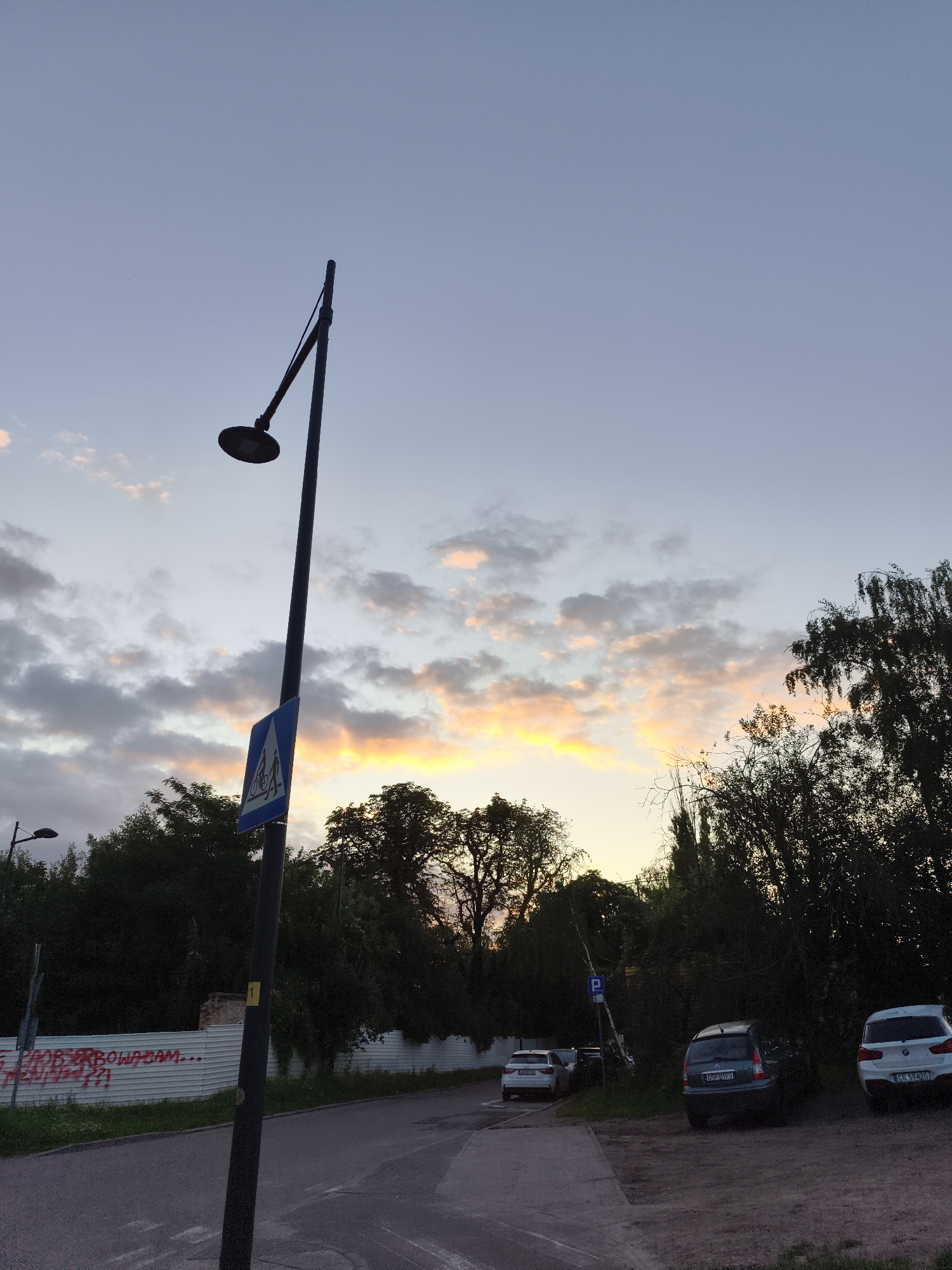 This is a low-angle, medium shot of a street scene at dusk in Gdańsk, Poland. The sky is a gradient of pale blue to soft orange and pink hues, with scattered clouds. A tall, dark lamppost with a curved arm stands prominently in the foreground, slightly off-center to the left. Attached to the lamppost is a blue, diamond-shaped traffic sign depicting a pedestrian and a cyclist crossing. To the left of the lamppost, a portion of a white fence with red graffiti is visible. Beyond the fence, trees and the silhouette of a building with a damaged brick wall can be seen. To the right of the lamppost, a paved road curves away. A few cars are parked on a dirt and gravel area to the right of the road, beneath a dense canopy of trees. The trees are dark and silhouetted against the fading light. One car, a dark blue hatchback, is in the middle ground, and two white SUVs are in the background. A blue parking sign is also visible near the parked cars. The overall atmosphere is quiet and subdued, indicative of the early evening hour.