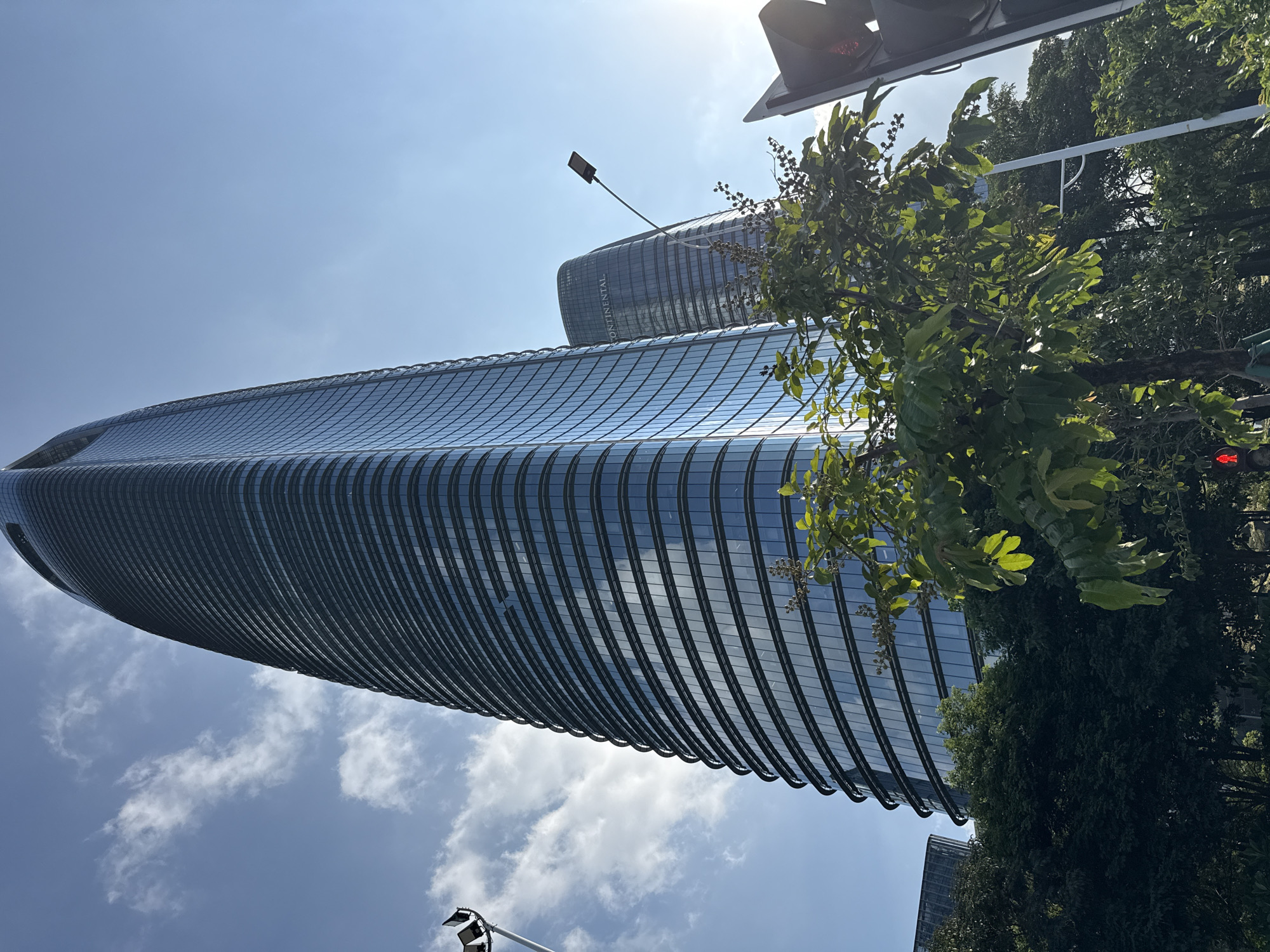 The image captures a low-angle view of a cluster of modern skyscrapers in Haizhu District, China. The foreground features lush green trees and foliage, with a glimpse of a traffic light and a pedestrian crossing signal. Dominating the frame is a sleek, curved skyscraper with a glass facade that reflects the bright blue sky and scattered clouds. To its right, another curved skyscraper is partially visible, bearing the "CONTINENTAL" logo. A street lamp with a modern design extends upwards towards the sky between the two prominent buildings. The overall scene suggests an urban environment with a blend of nature and contemporary architecture.