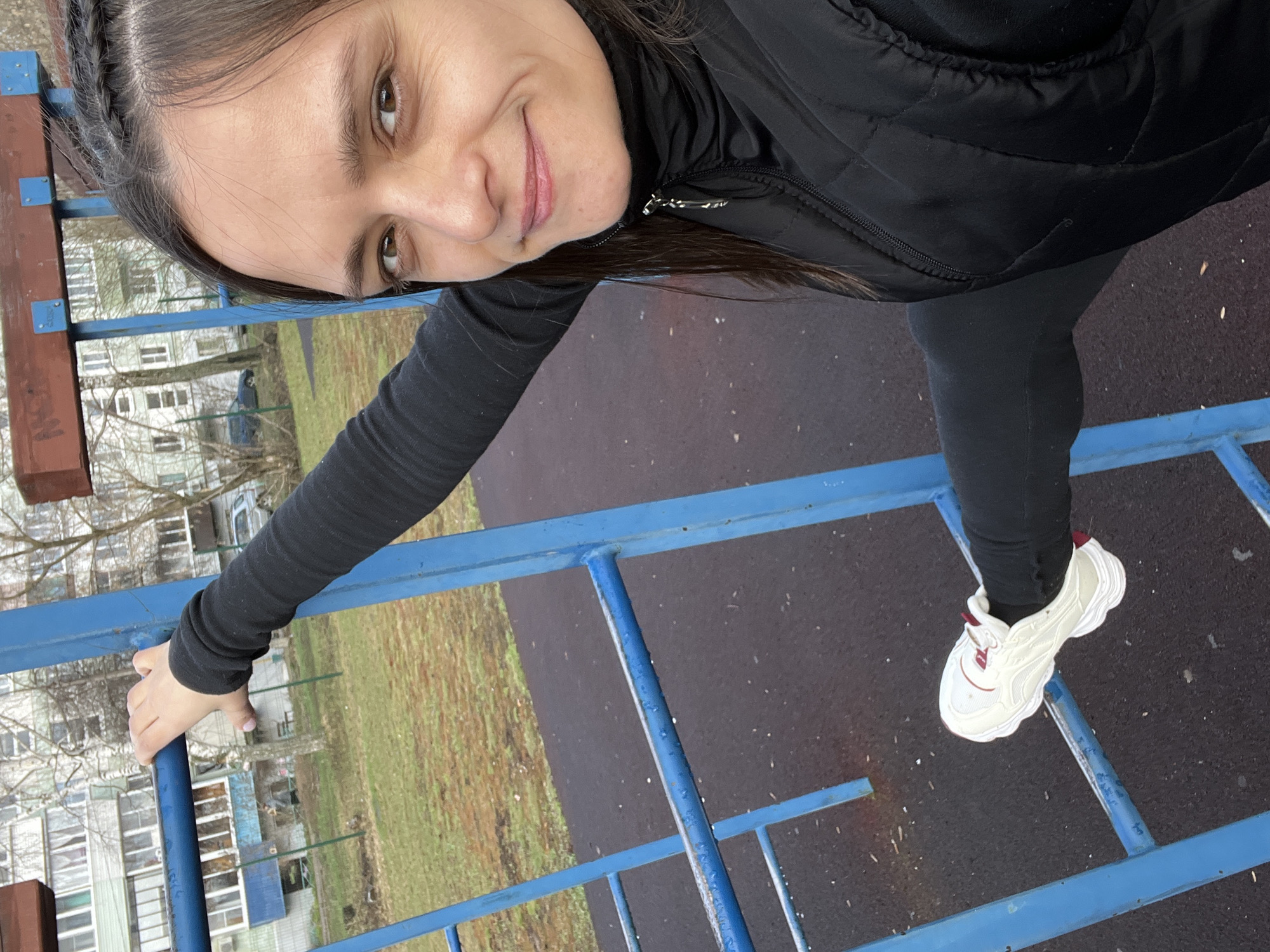 A young woman is depicted in an outdoor setting, posing on a blue metal climbing frame. She has dark hair, partially styled in braids near her forehead, and a faint smile on her face as she looks towards the camera. She is wearing a black long-sleeved top, a black quilted vest, black leggings, and white sneakers with subtle red accents. Her right hand grips a vertical blue bar of the climbing frame, while her left foot rests on a horizontal blue bar.
The background reveals a residential area with multi-story buildings, possibly apartment blocks, characterized by numerous windows. Bare trees without leaves are scattered throughout the scene, suggesting either late autumn, winter, or early spring. The ground is a mix of muted green grass/dirt and a dark, possibly asphalt or rubberized surface. A dimly visible figure in dark clothing can be seen in the distant background, near the buildings.
The lighting is soft and diffused, indicating an overcast day. Water droplets are visible on the blue metal bars of the climbing frame, suggesting damp conditions or recent rain. The woman's layered clothing also implies cool weather. Her expression appears calm and pleasant. There is no visible text in the image. The scene is set in Chaikovsky, Russia.