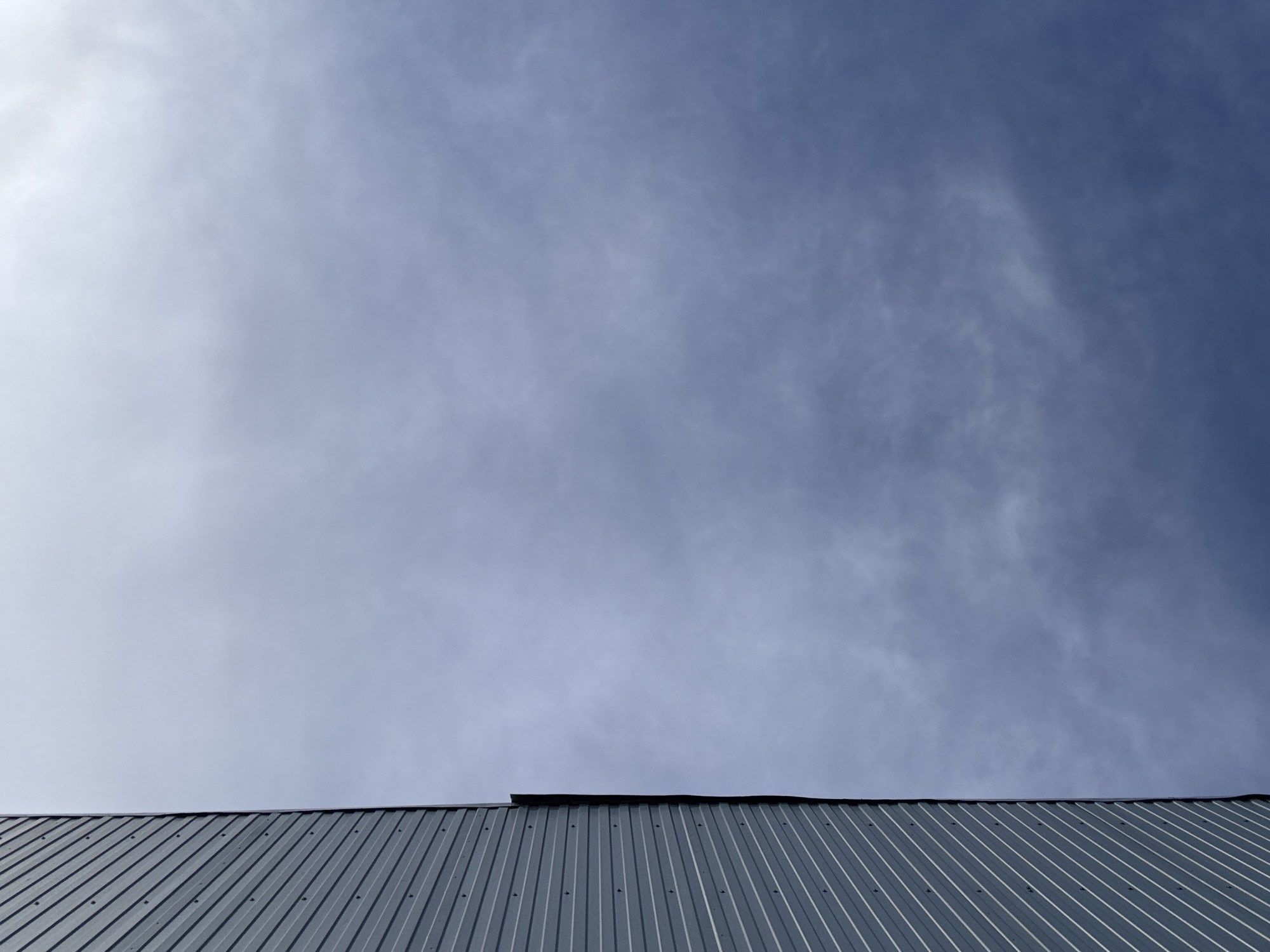 The image displays a view of a corrugated metal roof meeting a cloudy sky. The roof is metallic gray with visible fasteners along the ridges. The sky above is a gradient of blues and whites, with wispy clouds scattered across the expanse. The lighting suggests it is daytime, possibly overcast or during a transition in weather. The scene is devoid of any people or specific location markers beyond the roof itself. There is no visible text in the image.