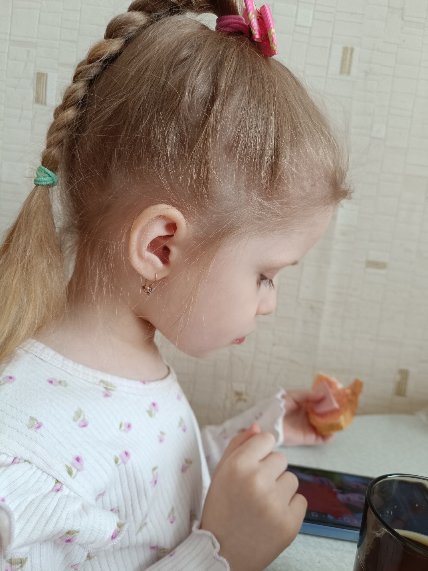 A young child with blonde hair braided on one side and secured with a pink hair clip is sitting indoors, likely at a table. The child wears a white long-sleeved shirt with a floral pattern. She is holding a piece of bread or pastry in one hand and looking down at a mobile phone or tablet on the table. A glass of liquid sits to the side of the device. The background features a light-colored wall with a subtle texture. Without additional information it is impossible to confirm location in Tobolsk, Russia.