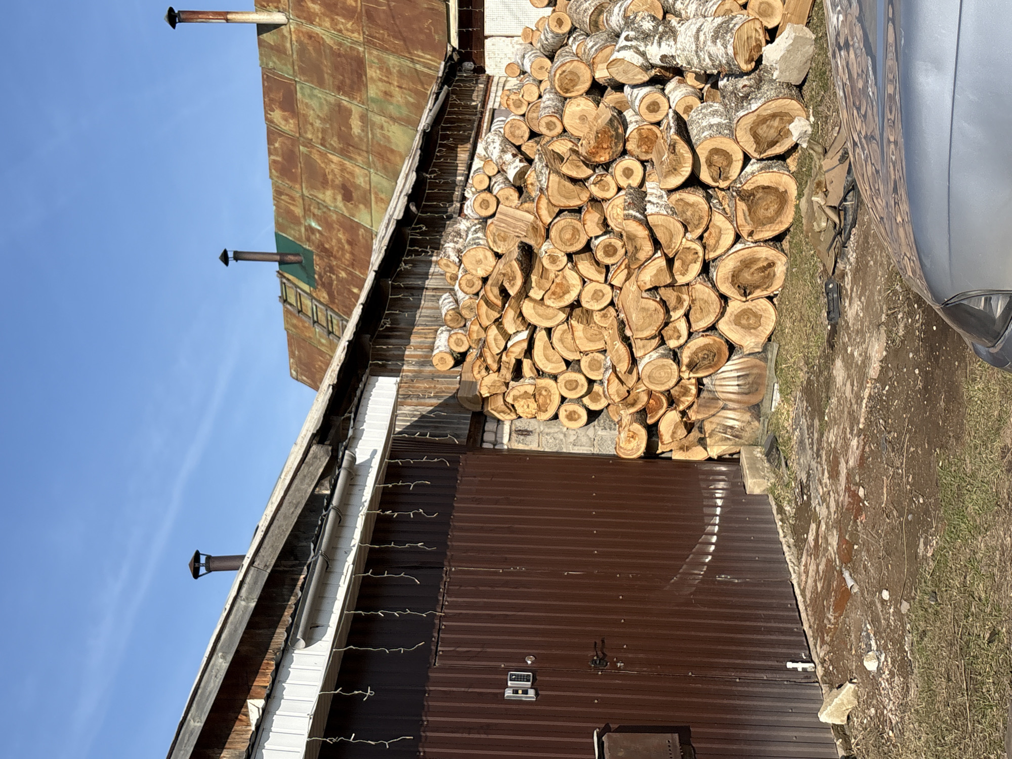 The image shows a stack of birch firewood next to a brown corrugated metal building in Chaikovsky, Russia. The building has a door and a security panel. Above, there is a white building with Christmas lights hung from its side. To the left, a green metal building with chimneys is visible against a blue sky. Some of the building is cut off by the right side of the frame. To the right of the firewood and corrugated metal building is a patch of dirt and grass. The front of a silver car is partially visible in the frame.