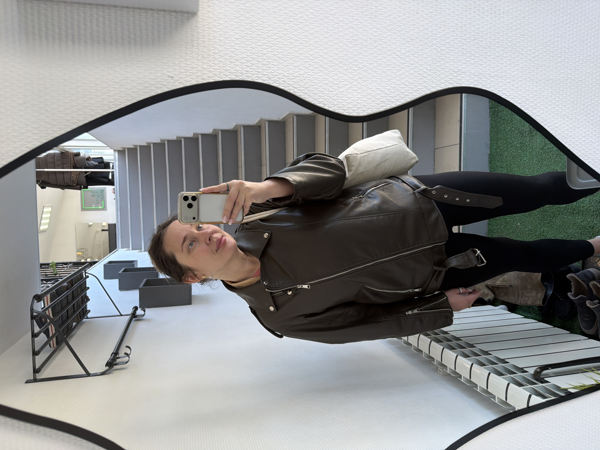 A mirror selfie shows a woman in a brown leather jacket and black leggings. She's holding a smartphone with her right hand, capturing the reflection. A white tote bag is slung over her shoulder. The mirror's frame is irregular, black, and wavy.
The reflection shows a hallway with a staircase and gray potted plants. A metal coat rack is visible on the left. The floor appears to be tiled in white. A green carpet or artificial turf is seen on the right edge of the reflection.
Based on previous knowledge, this is likely taken in a public space, such as a shop or business in the city of Chaikovsky, Russia.