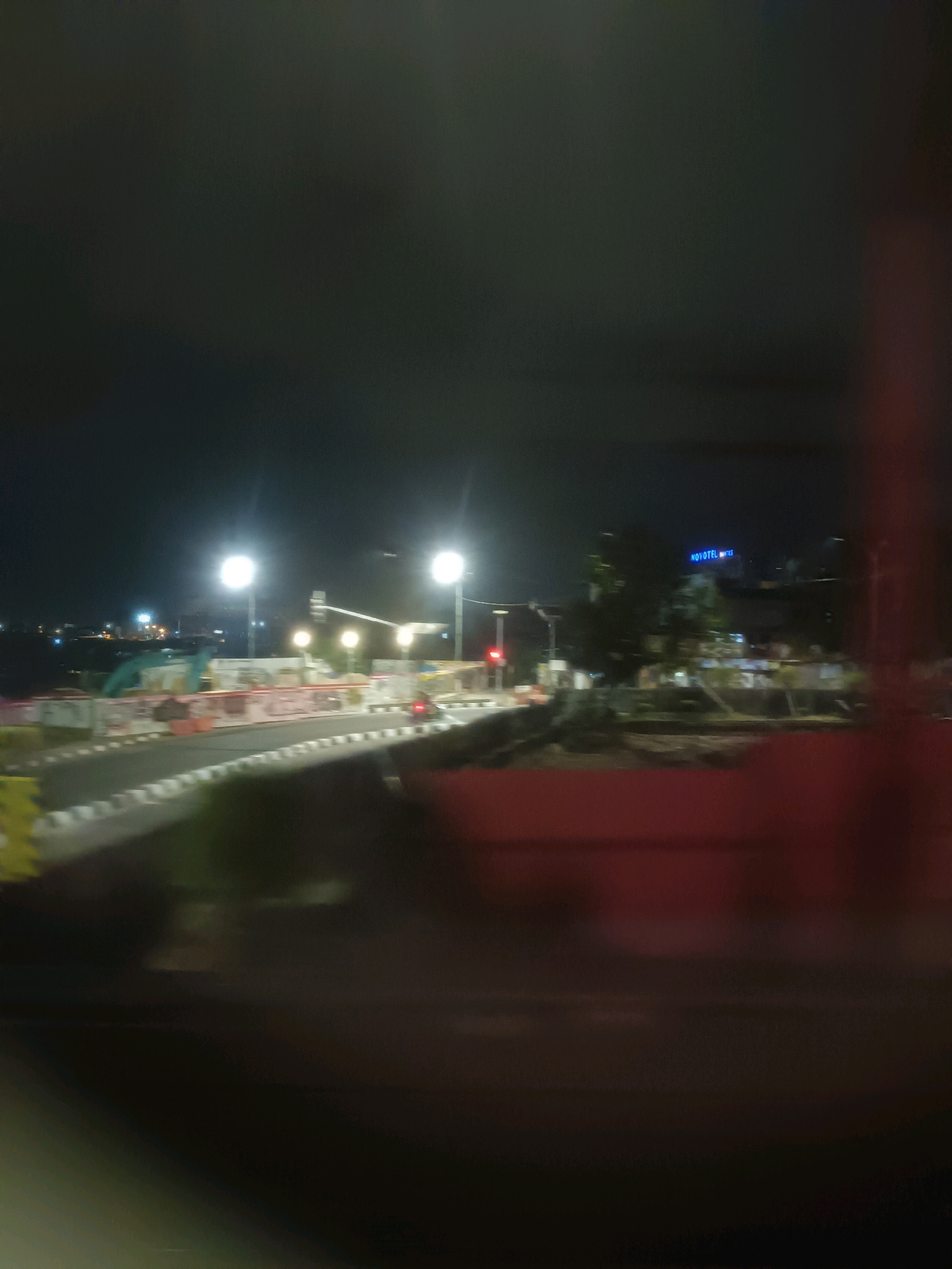 This is a nighttime, out-of-focus, exterior shot of a road under construction in Yogyakarta, Indonesia. The foreground features the road with white and black markings, and construction barriers. In the midground, there are several bright streetlights illuminating the area. Further back, a building with a prominent blue sign reading "NOVOTEL" is visible, suggesting the location is near a Novotel hotel. The scene is dark with minimal ambient light, indicating it is nighttime. There are no people or vehicles clearly identifiable due to the blurriness. The construction suggests ongoing development or maintenance in the area.