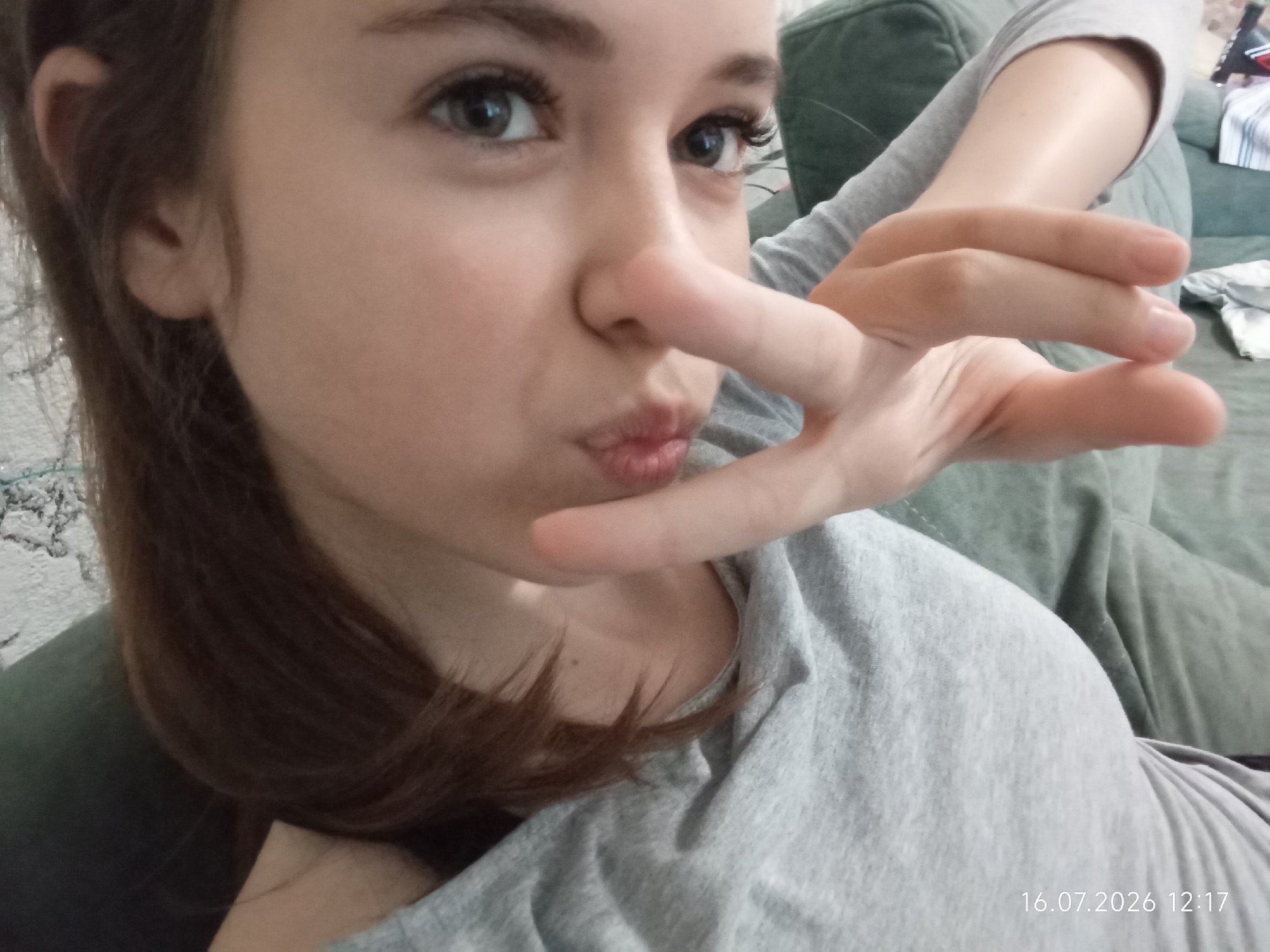 A close-up, selfie-style shot features a young person's face, with their hand obscuring part of their nose and mouth in a playful gesture. Their eyes are open and looking towards the camera, with long eyelashes visible. They are wearing a gray t-shirt. The background is blurred, showing a green sofa. A date and time stamp, "16.07.2026 12:17", is visible in the lower right corner. The image does not contain any information that would indicate its location within Qobda, Kazakhstan.