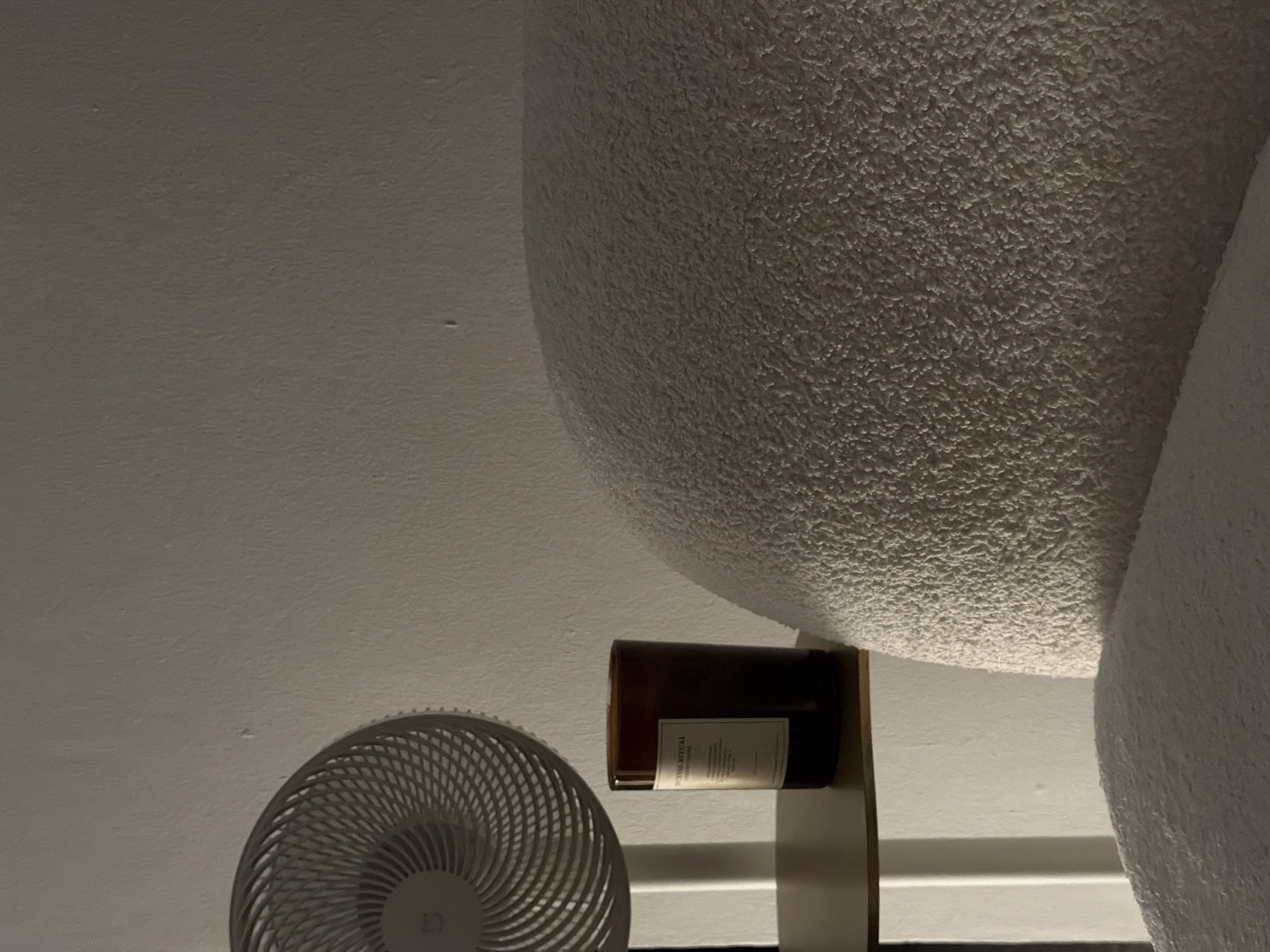 A fan, a candle, and a textured surface are depicted in this indoor scene. The fan, white with a circular grill pattern, is positioned on the left side of the image. To its right, a dark amber candle in a glass jar with a white label sits on a shelf. The dominant feature on the right side of the image is a plush, textured surface, possibly upholstery or fabric, which curves upwards and fills a significant portion of the frame. The background is a plain, lightly textured wall. The lighting is dim, creating soft shadows. The location is specified as Baan Plai Laem, Thailand.