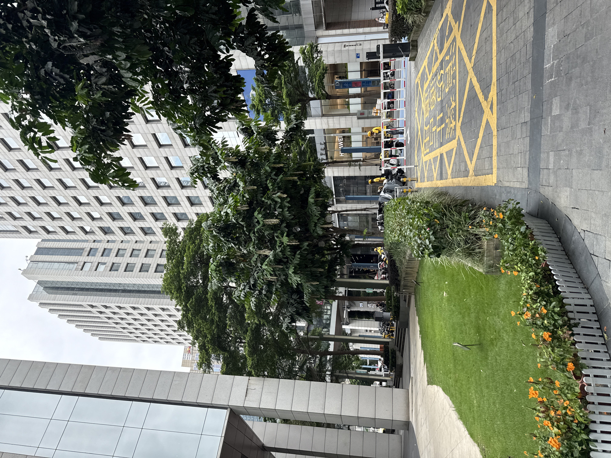 The image shows a street scene in Tianhe District, China, with tall modern buildings on either side. In the foreground, a patch of green grass is bordered by a white picket fence with blooming orange flowers. Paved pathways and a street with yellow markings are visible. Several trees with lush green foliage are scattered throughout the scene, with some branches extending over the buildings. In the background, there are parked motorcycles and scooters, and a white barrier with red and blue detailing across the street. A few individuals, some wearing yellow helmets, are present near the vehicles and barrier. The sky is overcast.