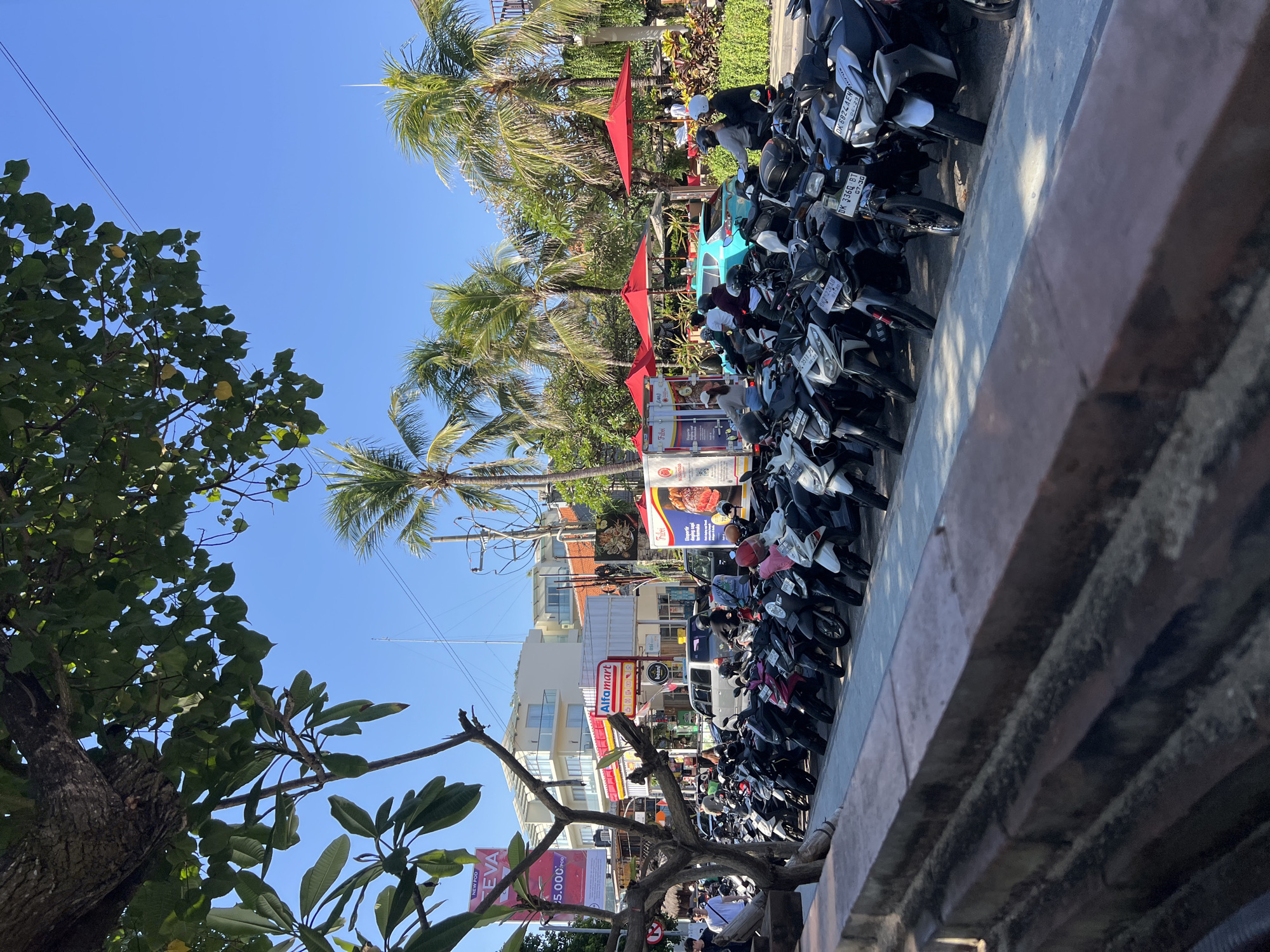 The image captures a street scene in Kuta, Indonesia, on a sunny day with a clear blue sky. In the foreground, a row of parked motorcycles, predominantly scooters, lines the paved area. Some motorcycles have helmets on their handlebars. Beyond the motorcycles, a sidewalk runs alongside a street.
On the other side of the street, a supermarket, "Alfamart," is visible with its red and white signage. Various shops and buildings are situated along the street, some with balconies. Palm trees are scattered throughout the scene, adding to the tropical ambiance. Red umbrellas are set up, suggesting an outdoor seating area for a cafe or restaurant.
Several people are present. Some are walking, while others appear to be on or near motorcycles, possibly preparing to depart or having just arrived. A light blue car is parked on the street. The overall impression is of a bustling commercial area in a tourist-oriented part of Kuta.