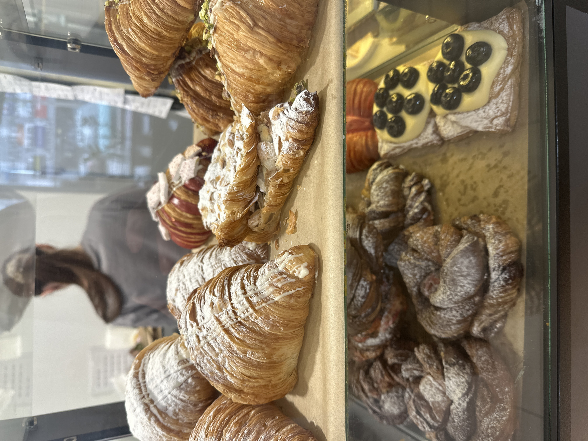 The image shows a display case in what appears to be a bakery or pastry shop. The display case contains a variety of pastries, including several croissants with powdered sugar, a croissant with pistachio nuts, another with stripes of icing and a pastry topped with custard and blueberries. A person is reflected in the glass of the display case. The location is not possible to determine from this image.